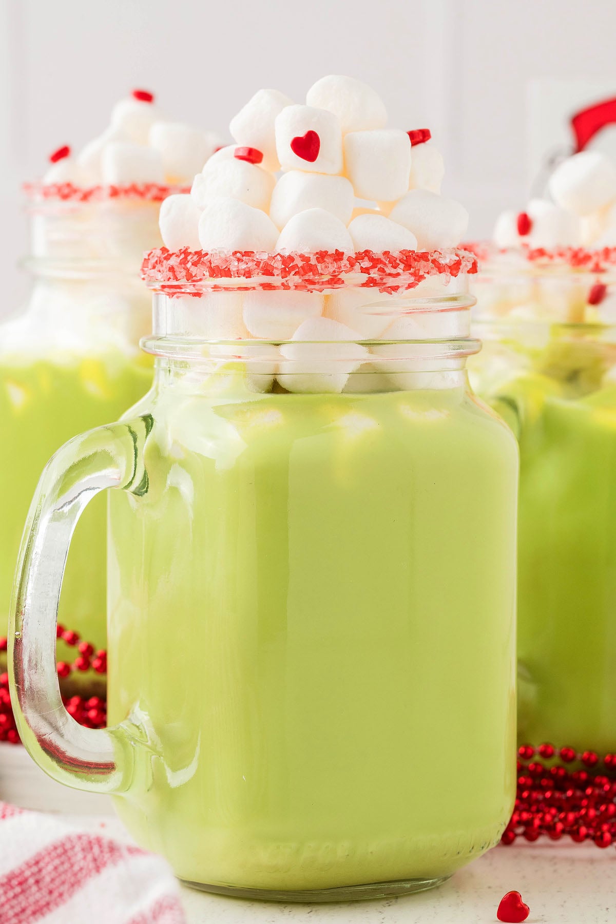 A green Grinch hot chocolate in a mason jar topped with marshmallows and red candy hearts on the rim—perfect as a fun holiday party drink.