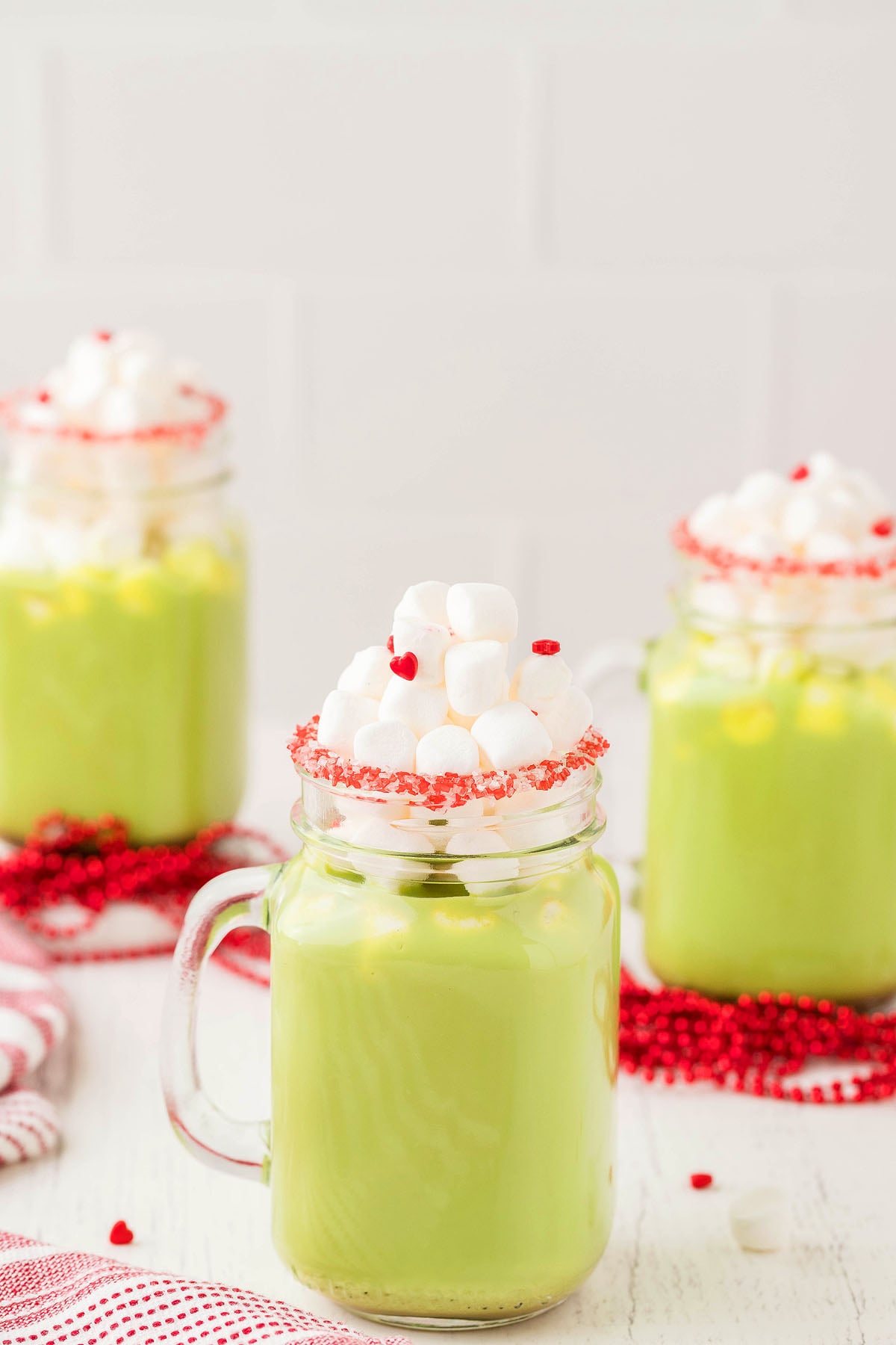 Three mugs of Green Grinch hot chocolate topped with mini marshmallows and red sprinkles, on a white surface—perfect as a Fun Holiday Party Drink.