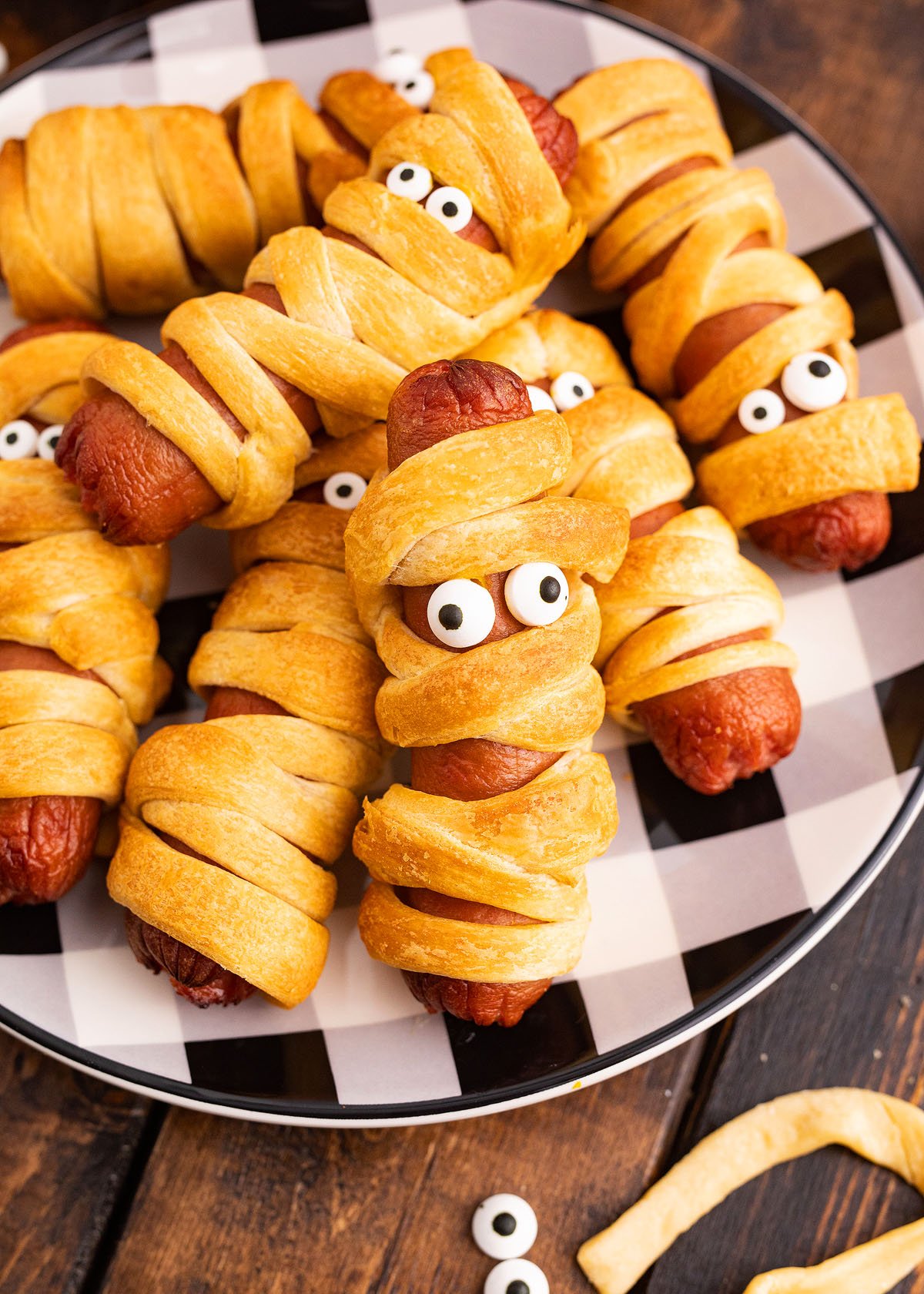 Crescent roll hot dog mummies are spooky and fun—scary hot dogs wrapped to look like mummies, complete with candy eyes, served on a black and white plate for the perfect Halloween treat.