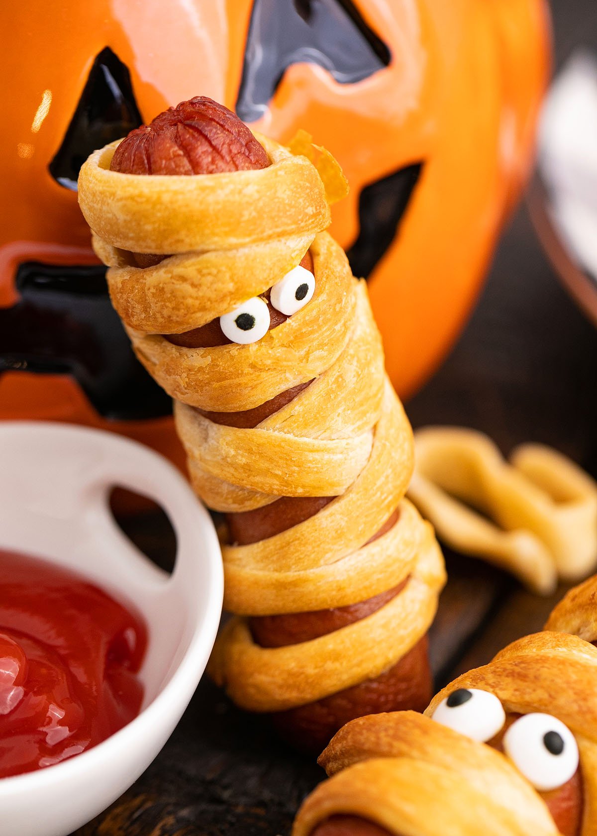 A scary hot dog wrapped in pastry like a mummy, complete with candy eyes, sits beside ketchup and a jack-o’-lantern—perfect as a Mummy Halloween Hot Dogs treat for your next Halloween party recipe!.