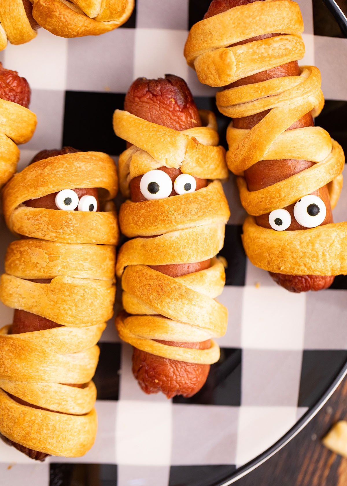 Three scary hot dogs wrapped in crescent roll dough to look like spooky pigs in a blanket mummies, complete with candy eyes, served on a checkered plate—a perfect Halloween party recipe.