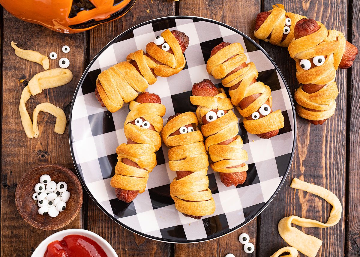 Spooky pigs in a blanket wrapped like mummies with candy eyes on a plaid plate, set against a Halloween-themed background.