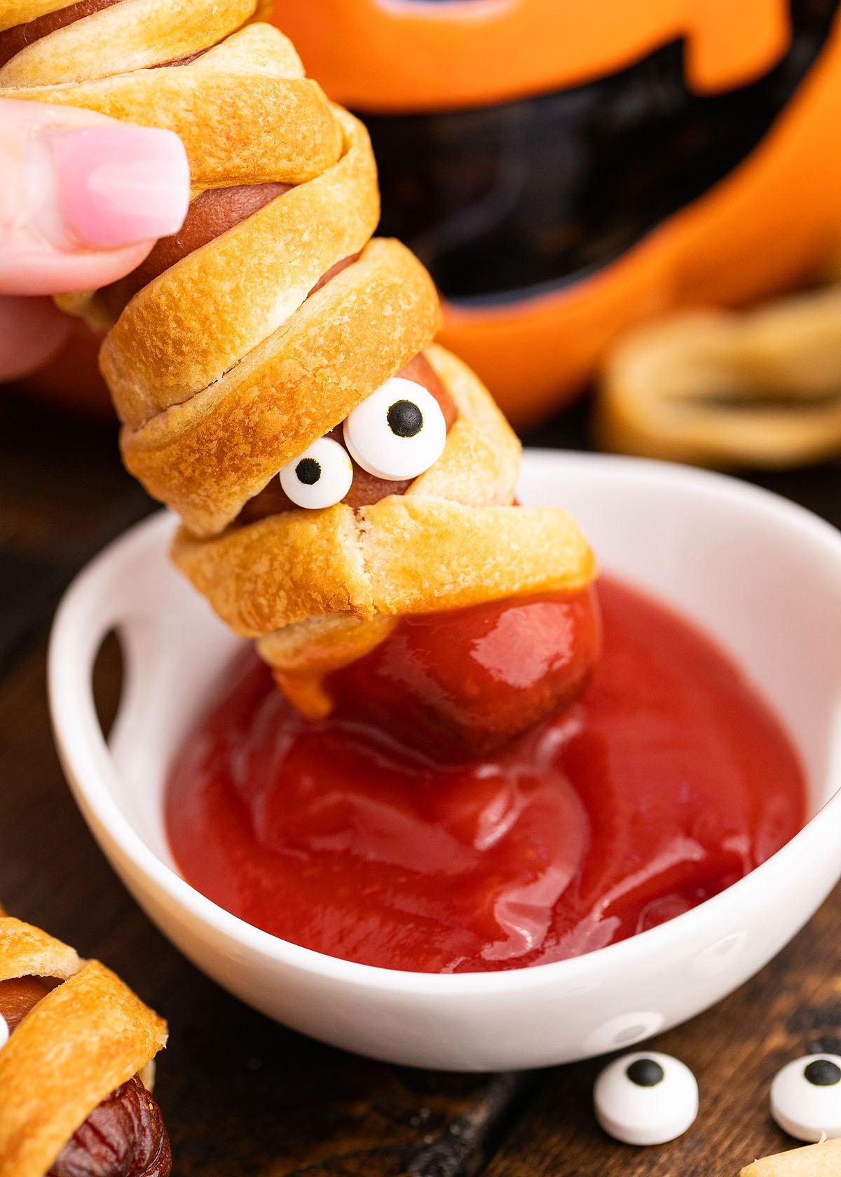 A sausage wrapped in dough like a mummy, with candy eyes, being dipped in ketchup—these spooky pigs in a blanket make the perfect Halloween party recipe.