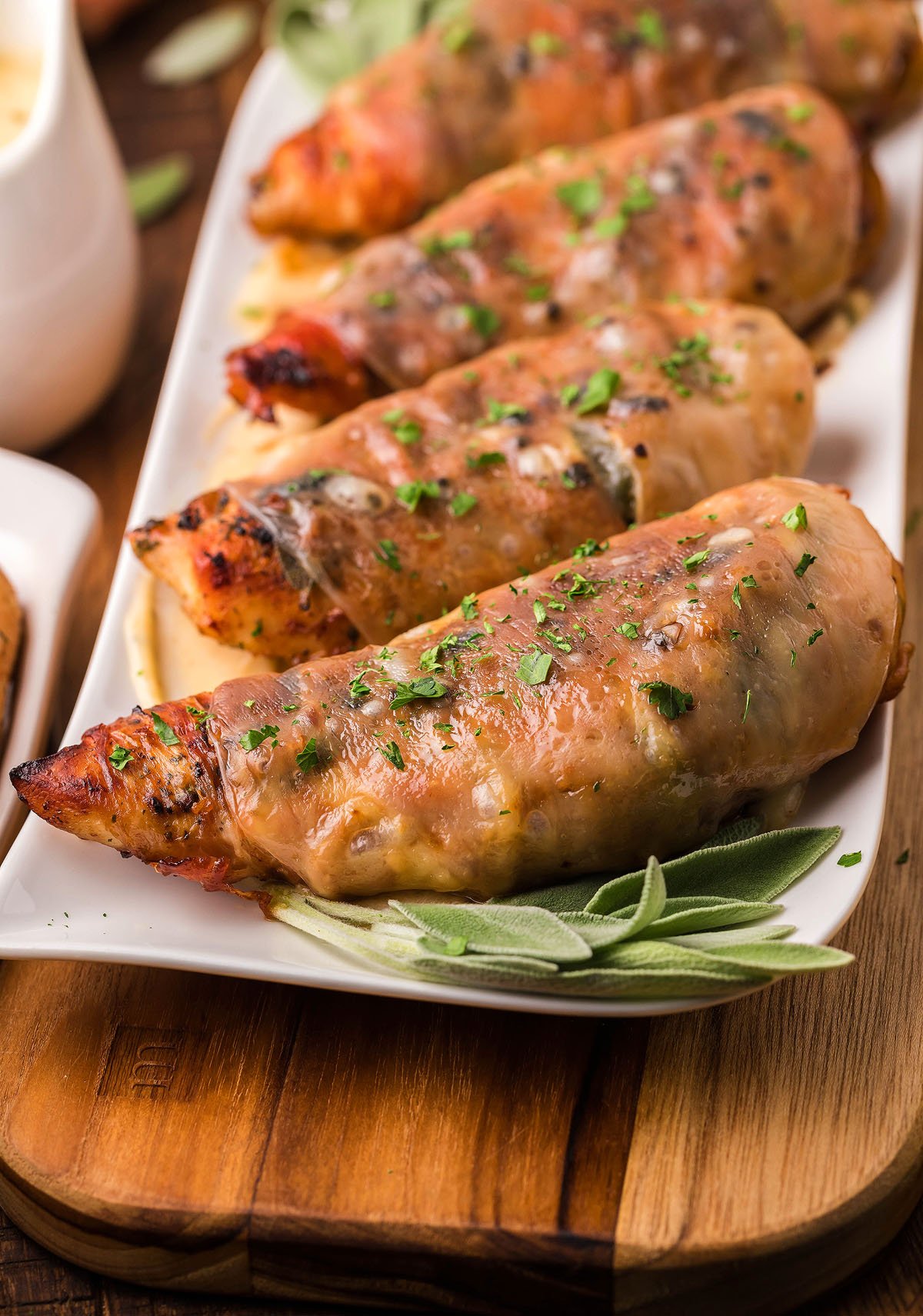 Four Chicken Saltimbocca breasts wrapped in prosciutto and garnished with herbs on a white platter with fresh sage leaves—an Italian baked chicken classic.