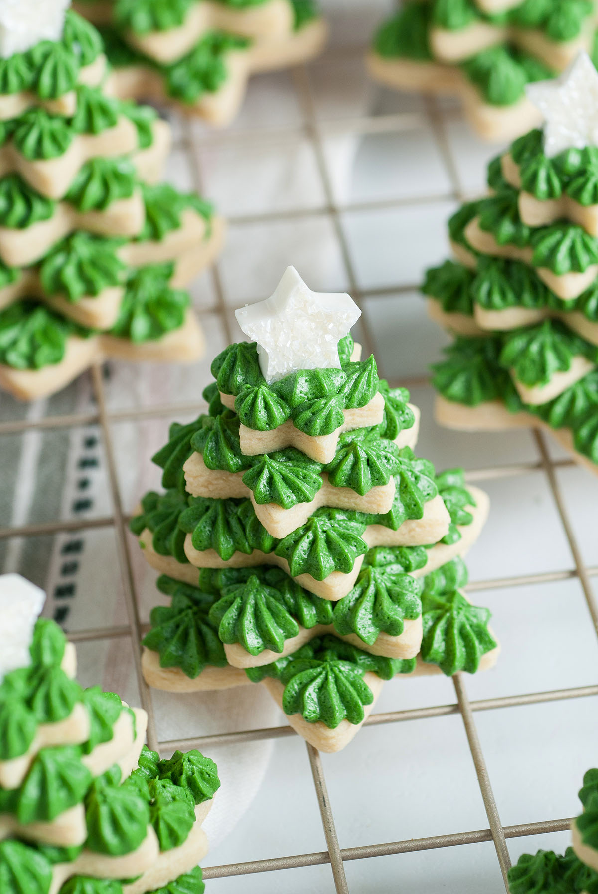 Stacked cookie trees in festive tree shapes, topped with green icing and a white star made with star cookie cutters, displayed on a cooling rack.