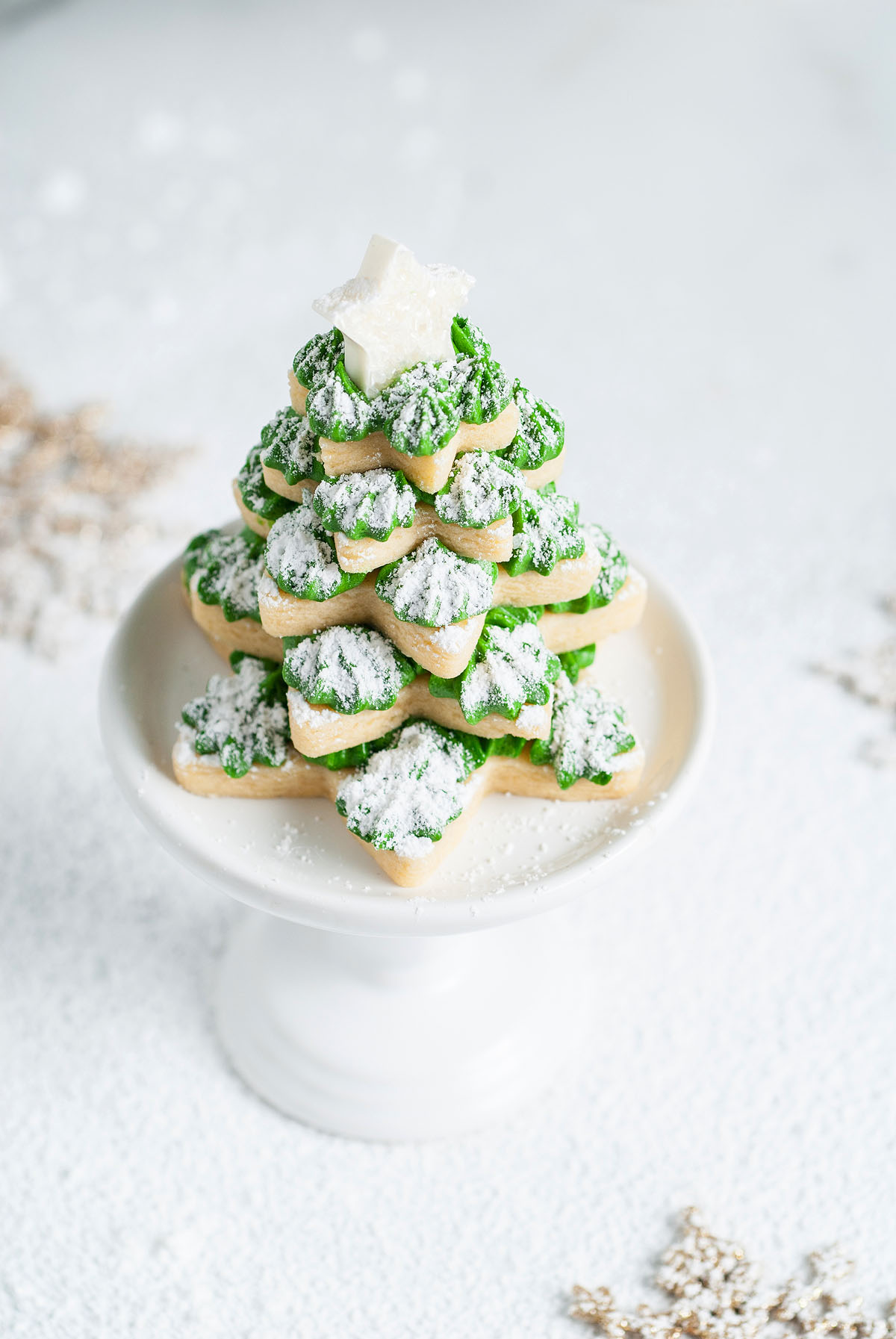 Stacked star-shaped cookies, made with star cookie cutters and green icing, form a festive Christmas tree topped with a white star, displayed on a white stand.