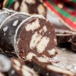 No bake Italian Chocolate Salami – Salame di Cioccolato, a sliced chocolate log with white cookie pieces, dusted with powdered sugar, sits in front of festive cloth.