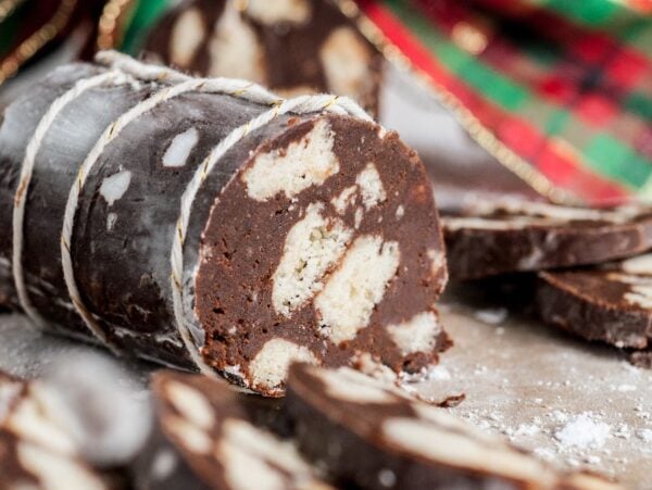 No bake Italian Chocolate Salami – Salame di Cioccolato, a sliced chocolate log with white cookie pieces, dusted with powdered sugar, sits in front of festive cloth.