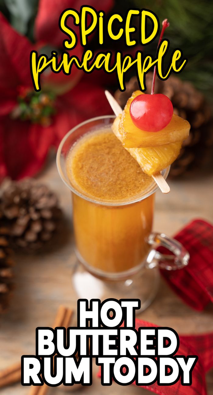 A glass of warm rum drink—this Rum Toddy blends black spiced rum and butterscotch schnapps, garnished with pineapple and a cherry, surrounded by festive decor.