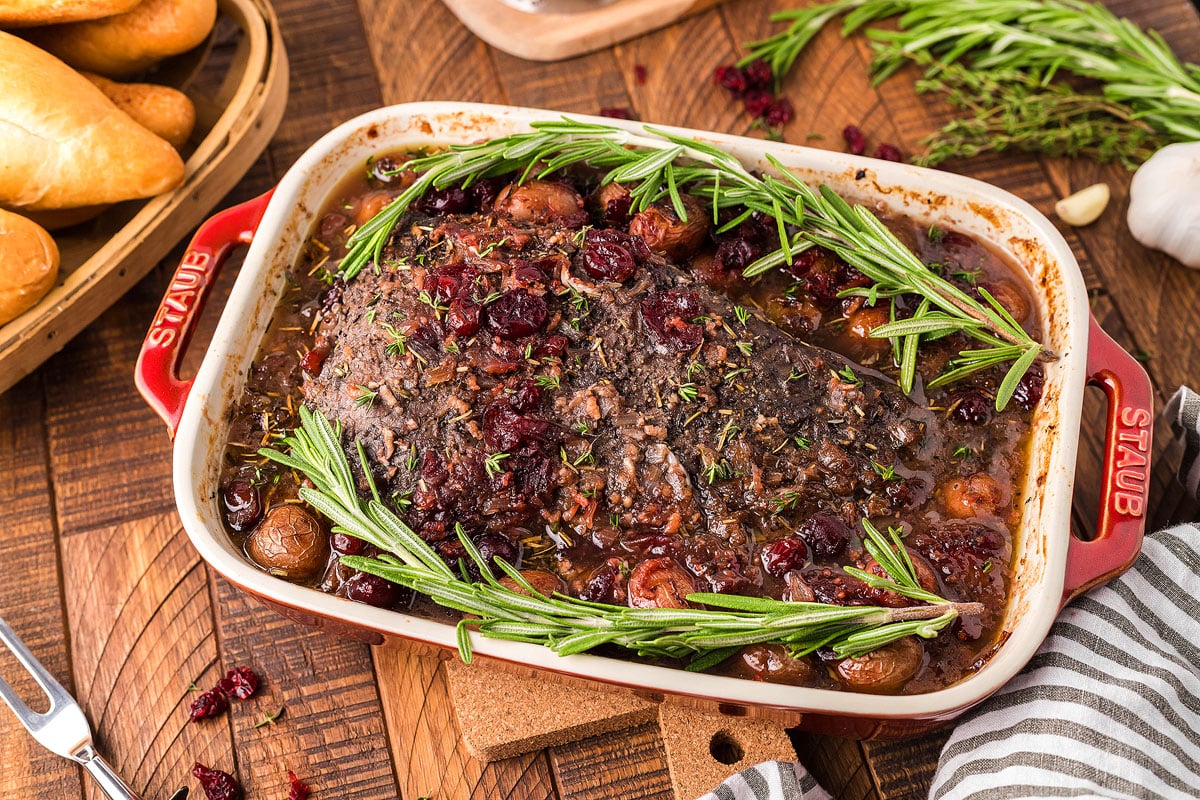 A baked dish in a red Staub pan contains a large roast covered in herbs, surrounded by sprigs of rosemary. The pan sits on a wooden table near a basket of bread rolls. Fresh garlic cloves and additional sprigs of rosemary are visible in the background.