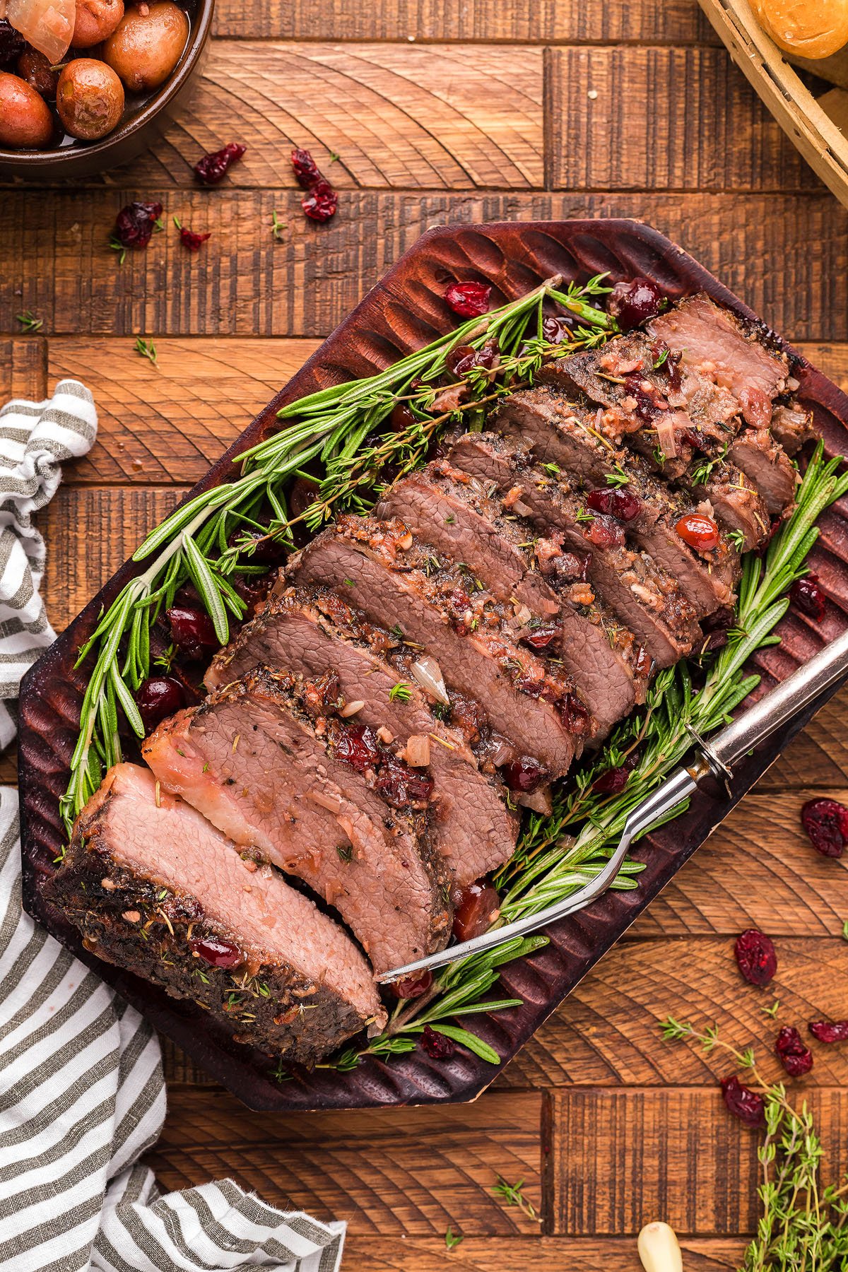 Sliced roast beef garnished with rosemary on a platter, perfect for a holiday meal—complete with a fork and striped napkin nearby.
