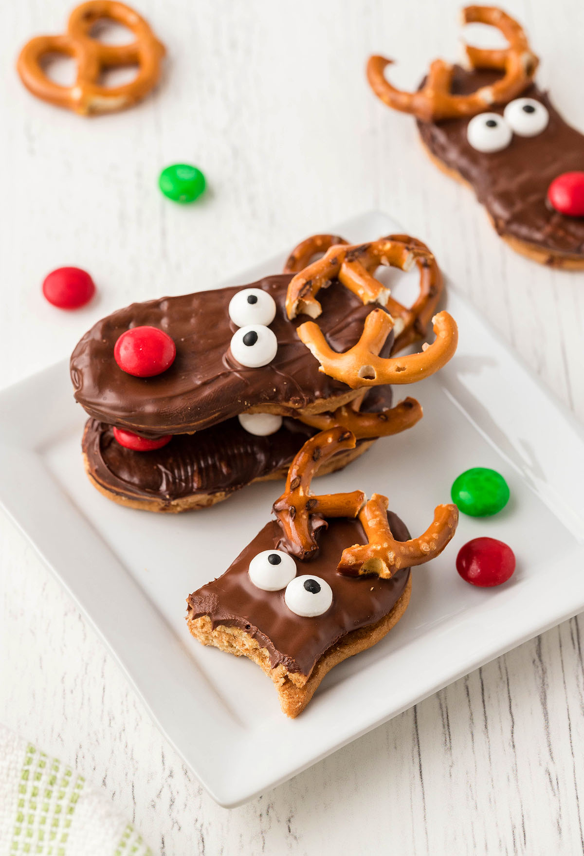 Three Nutter Butter Reindeer cookies with pretzel antlers and candy eyes sit on a white plate, decorated with red and green candies—perfect Christmas treats for holiday parties.