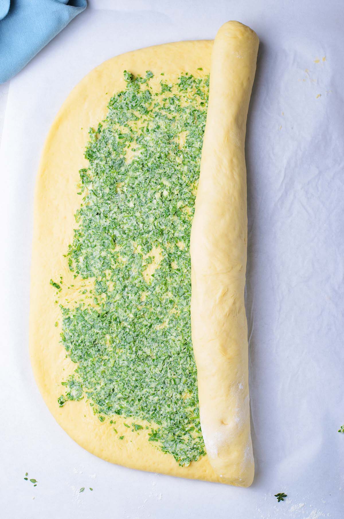 Dough for an easy bread recipe is topped with a green herb mixture and rolled up on a parchment-lined surface, perfect for making a flavorful garlic herb bread wreath.
