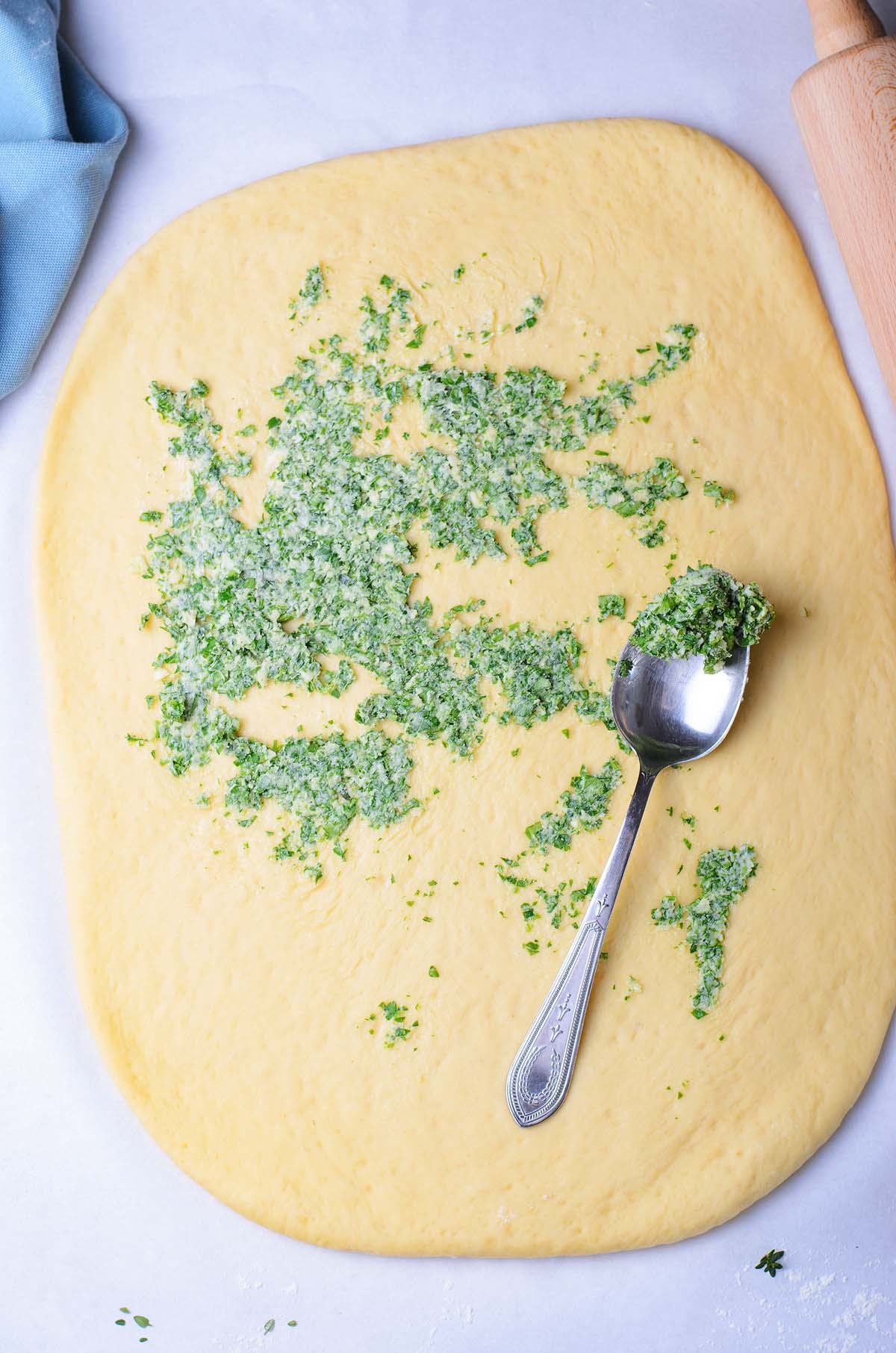 Rolled dough topped with a green herb mixture, perfect for making homemade bread or garlic bread, with a spoon and rolling pin on the side.