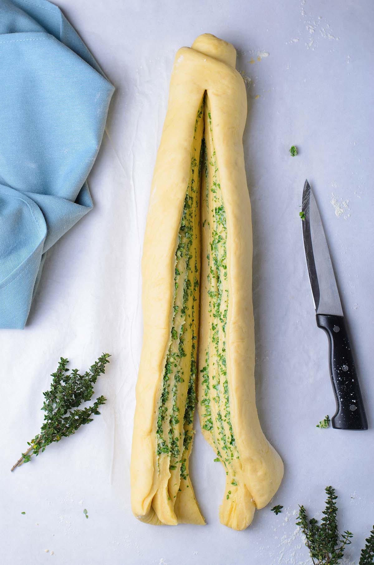 Dough for this easy bread recipe is filled with green herbs and sliced lengthwise, with a knife, thyme, and blue cloth nearby.