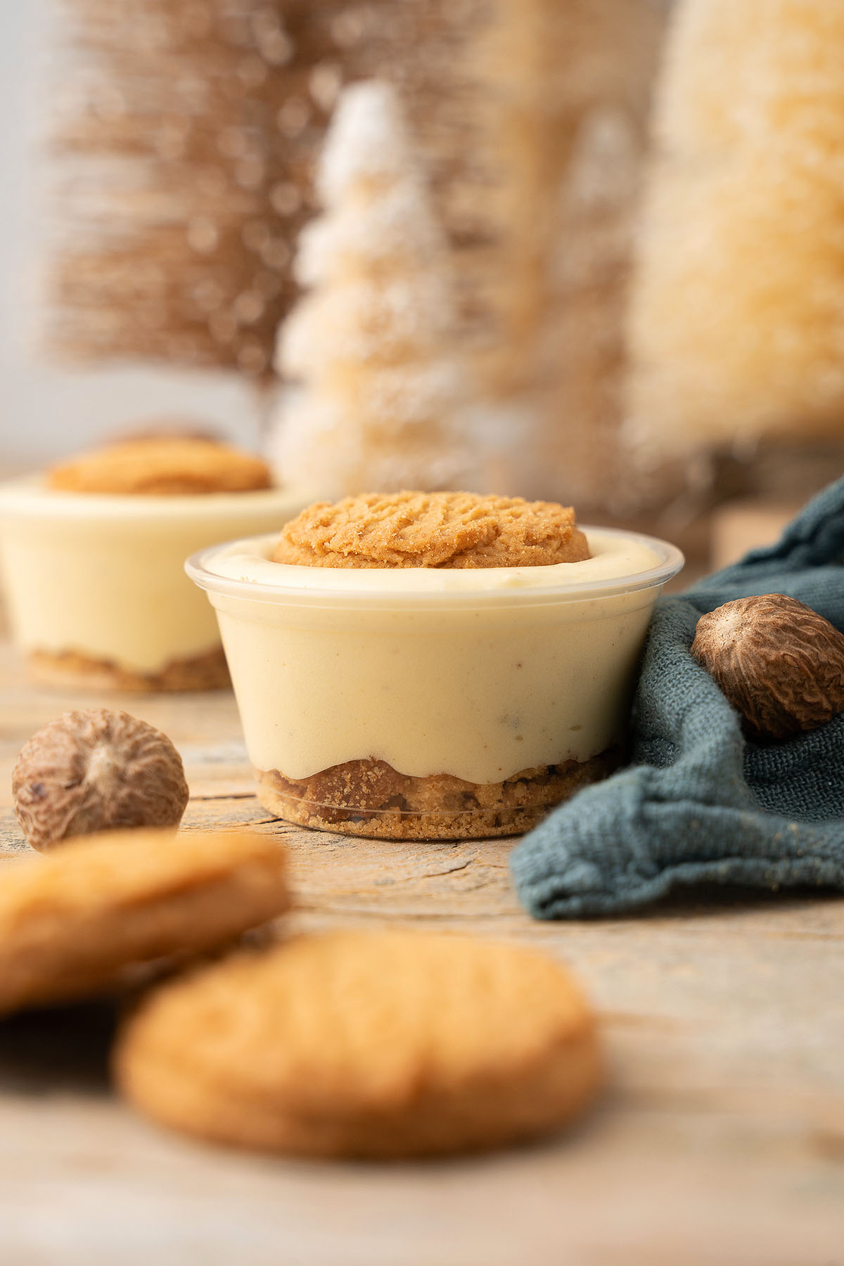 Mini cheesecake in a clear cup topped with a cookie, surrounded by spices and cookies on a wooden surface—perfect for fans of boozy pudding cups.