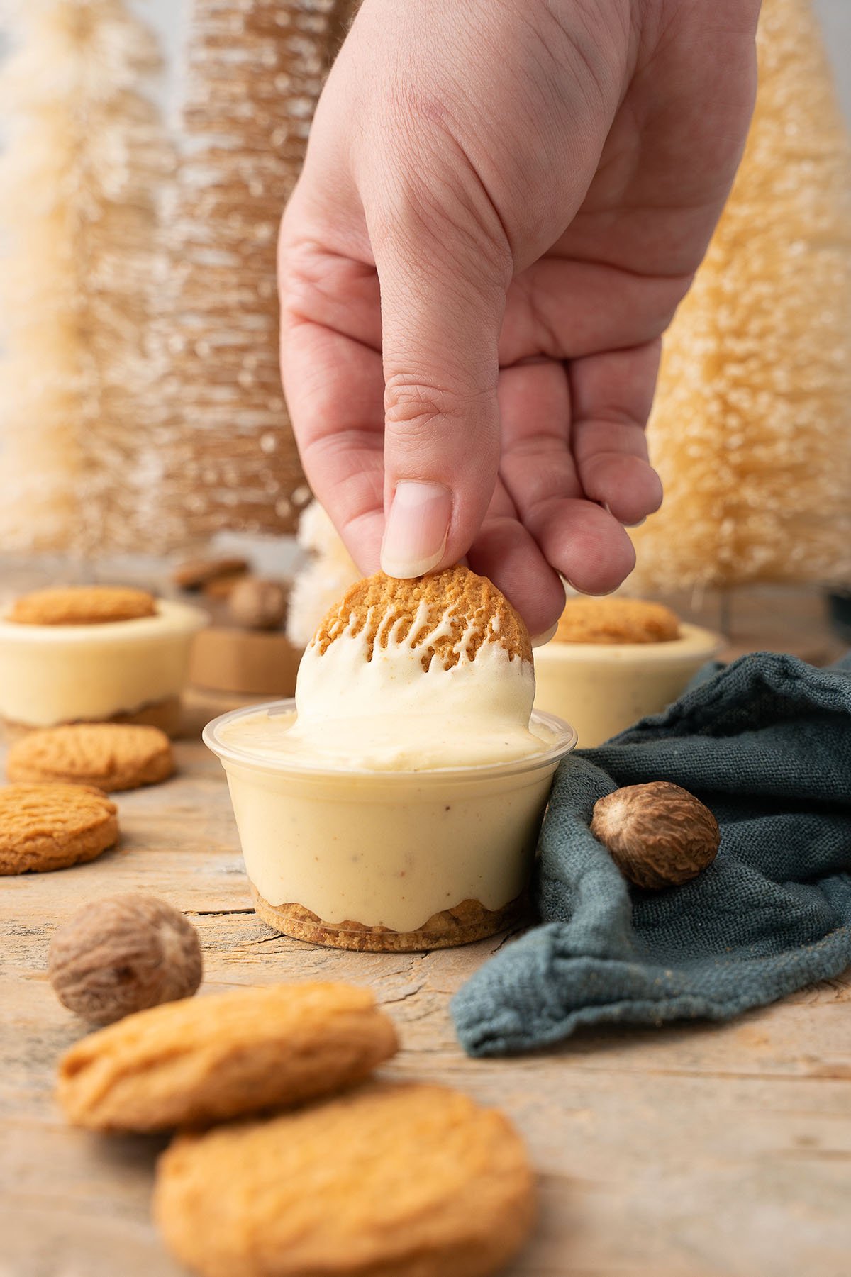 A hand dips a cookie into a small cup of creamy Ginger Snap Eggnog Pudding Shot, with more cookies, whole nutmeg, and hints of Black spiced rum and RumChata nearby—perfect for Christmas or holiday pudding shots.