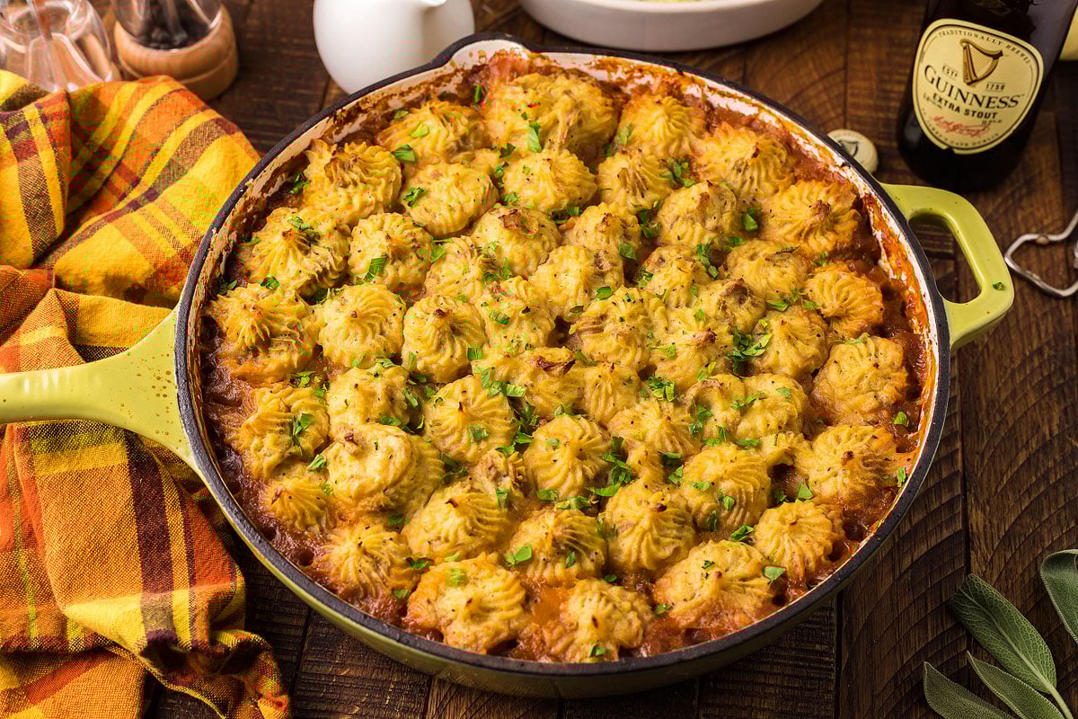 A round green dish filled with golden brown mashed potato-topped shepherd's pie. The dish is garnished with chopped herbs. A colorful plaid cloth is to the left, and a bottle of Guinness beer is on the right. Various kitchen items are in the background.