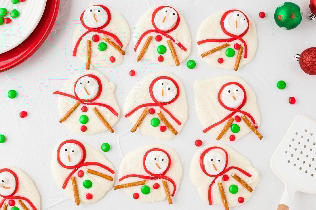 Snowman sugar cookies decorated with red icing, pretzel arms, and green and red candies, resembling melting snowman treats on a white background.