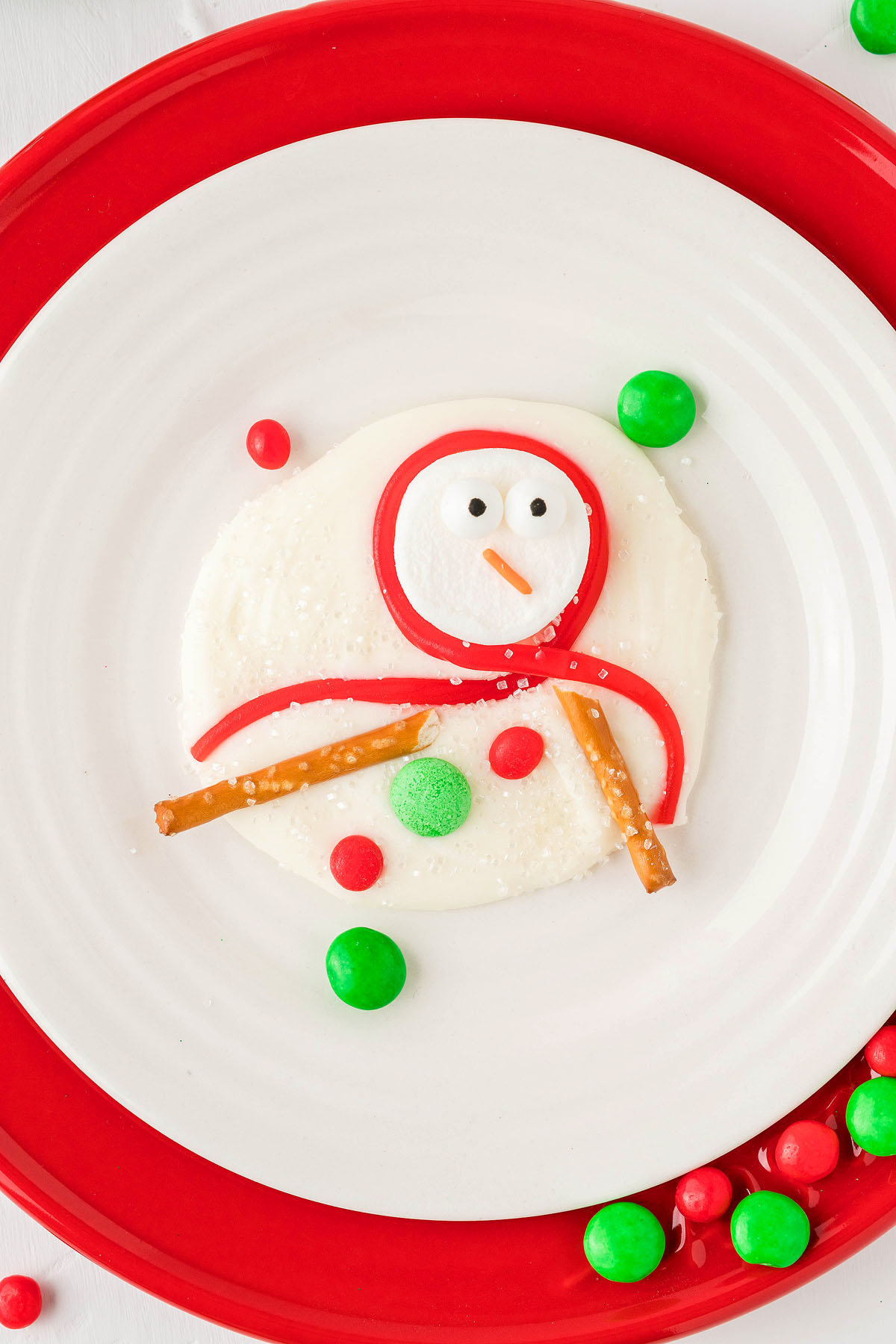 A snowman treat made of white chocolate Christmas candy, pretzel arms, red and green candies, and a marshmallow head on a plate.
