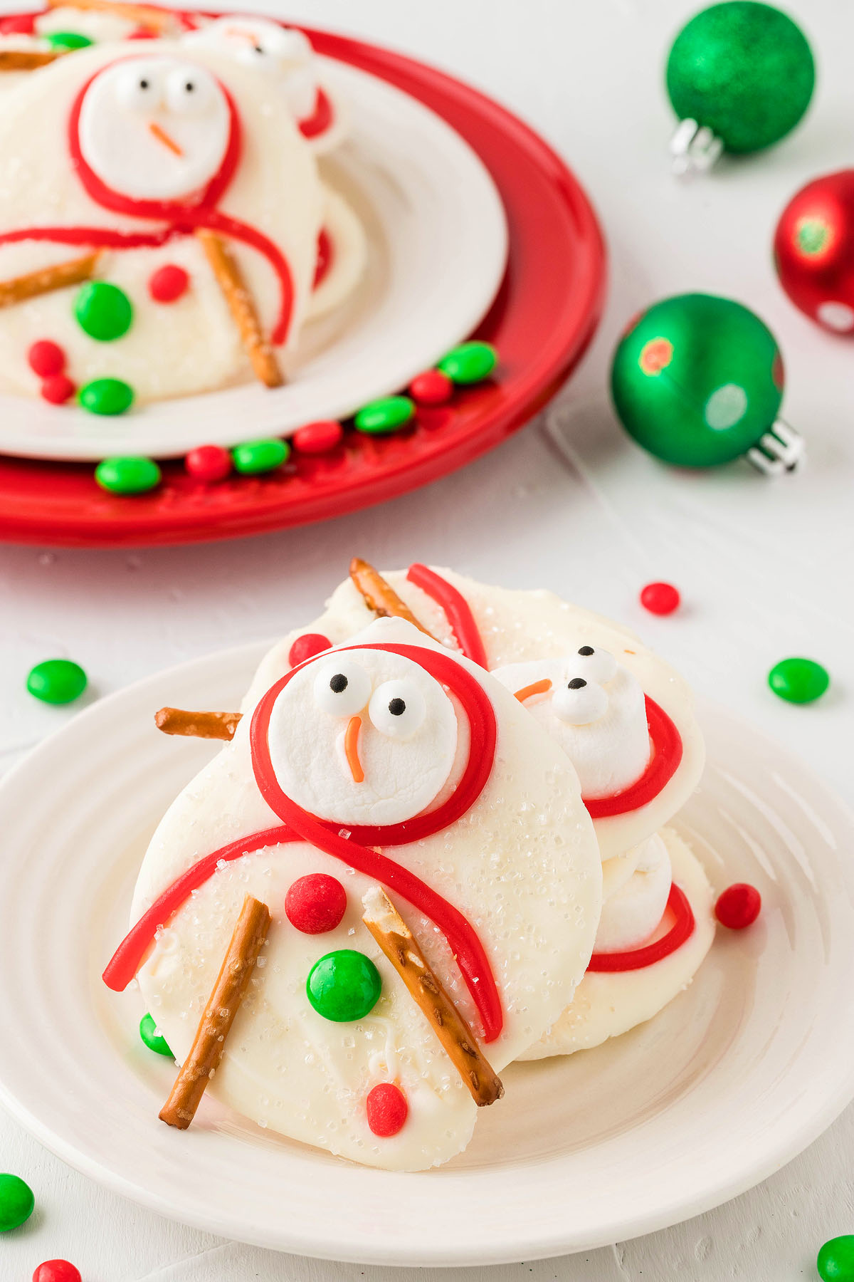 White chocolate Melted Snowman Cookie Bark with pretzel arms and candy decorations on a plate, surrounded by Christmas ornaments—a fun melting snowman treat perfect for holiday parties.