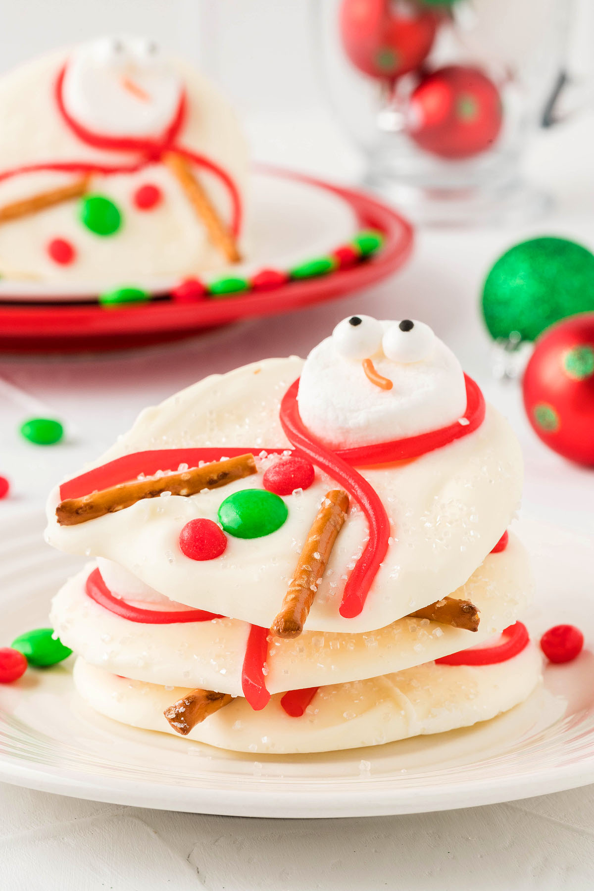 Stacked snowman cookies decorated with marshmallows, pretzel sticks, and colorful red and green candies capture the fun of Melted Snowman Cookie Bark—perfect as a whimsical white chocolate Christmas candy or festive Winter bark treat.