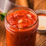 A glass jar filled with classic marinara sits on a wooden table beside its lid, showcasing the rich texture of this homemade Italian marinara sauce recipe.