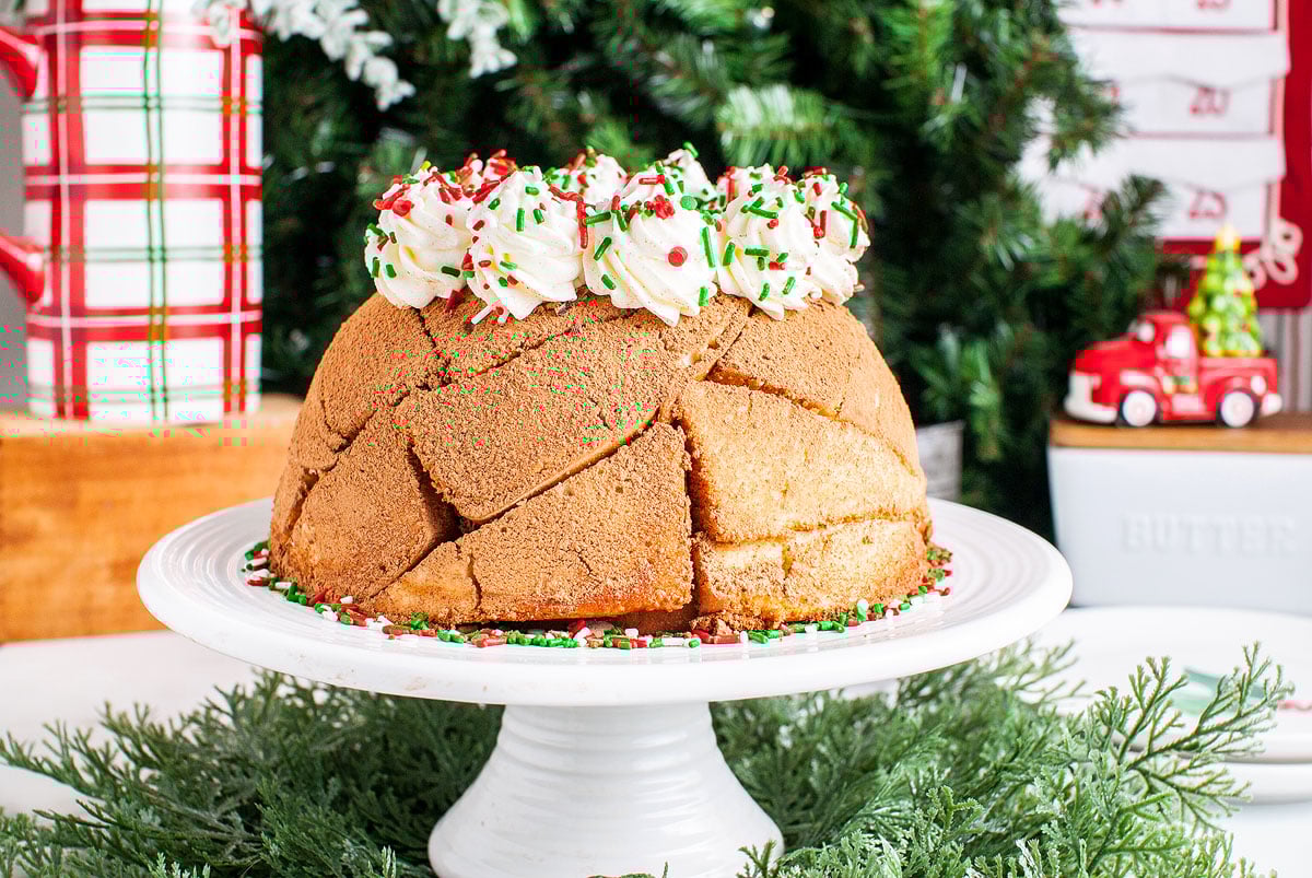 A festive Italian Christmas Zuccotto Cake with whipped cream and sprinkles sits on a white stand, surrounded by Christmas decor—an irresistible classic among Italian Christmas desserts.