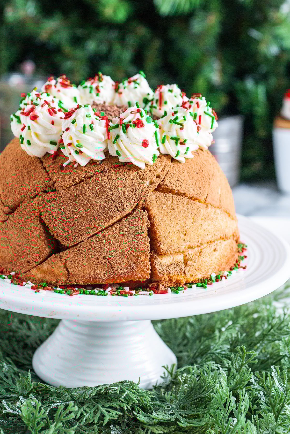 A round Italian Christmas Zuccotto Cake topped with whipped cream and red and green sprinkles sits on a white cake stand, perfect for holiday celebrations.
