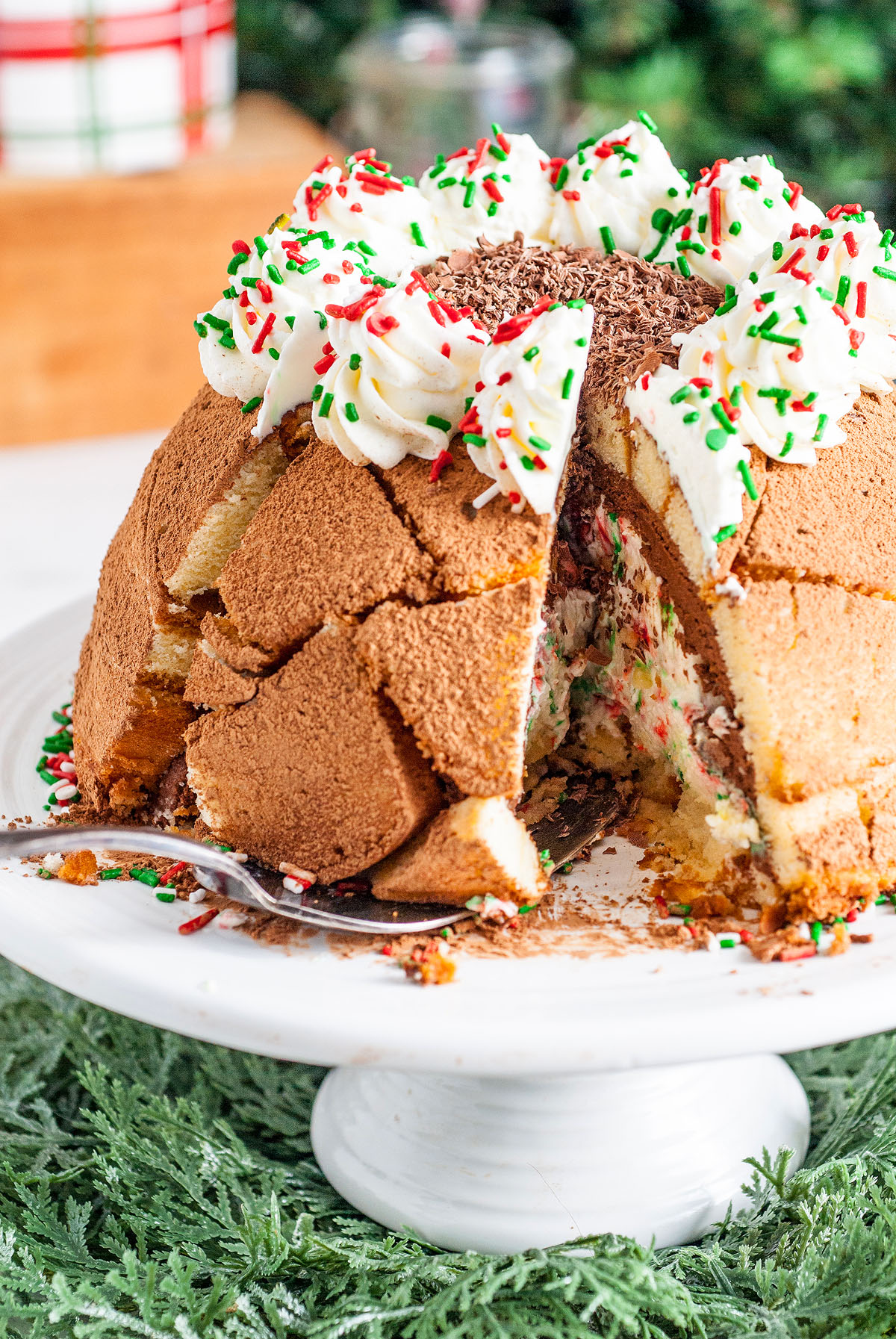 A festive dome-shaped Italian Christmas Zuccotto Cake with whipped cream, sprinkles, and a slice cut out, displayed on a white cake stand.
