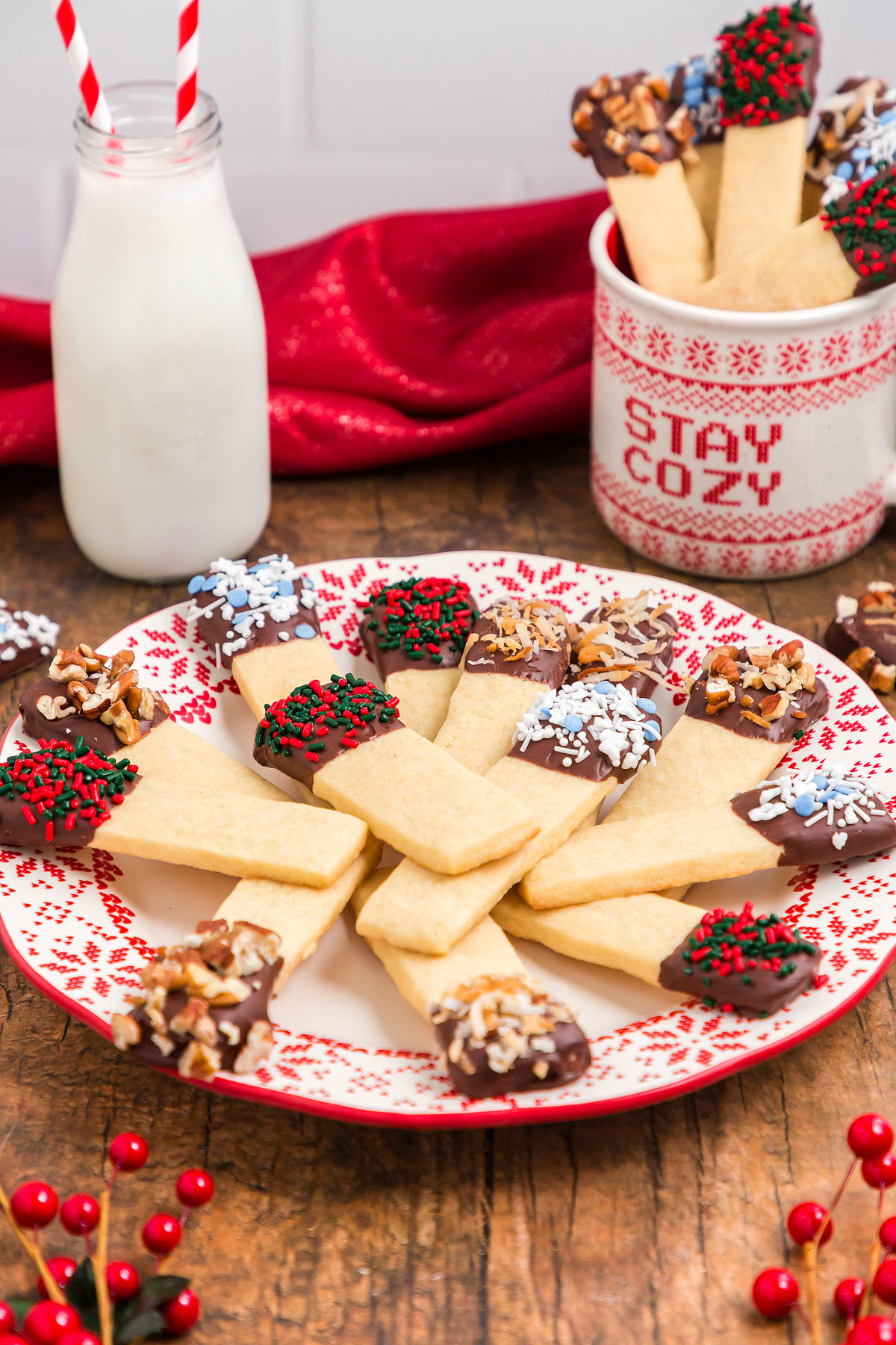 Plate of chocolate dipped cookie sticks with festive sprinkles, perfect for a cookie exchange, next to milk and a mug that says &ldquo;Stay Cozy.&rdquo;.