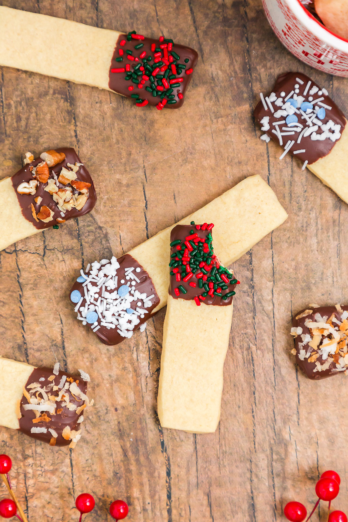Rectangular Dipped Sugar Cookie Sticks are dipped in chocolate and topped with colorful sprinkles, nuts, and coconut on a wooden table.