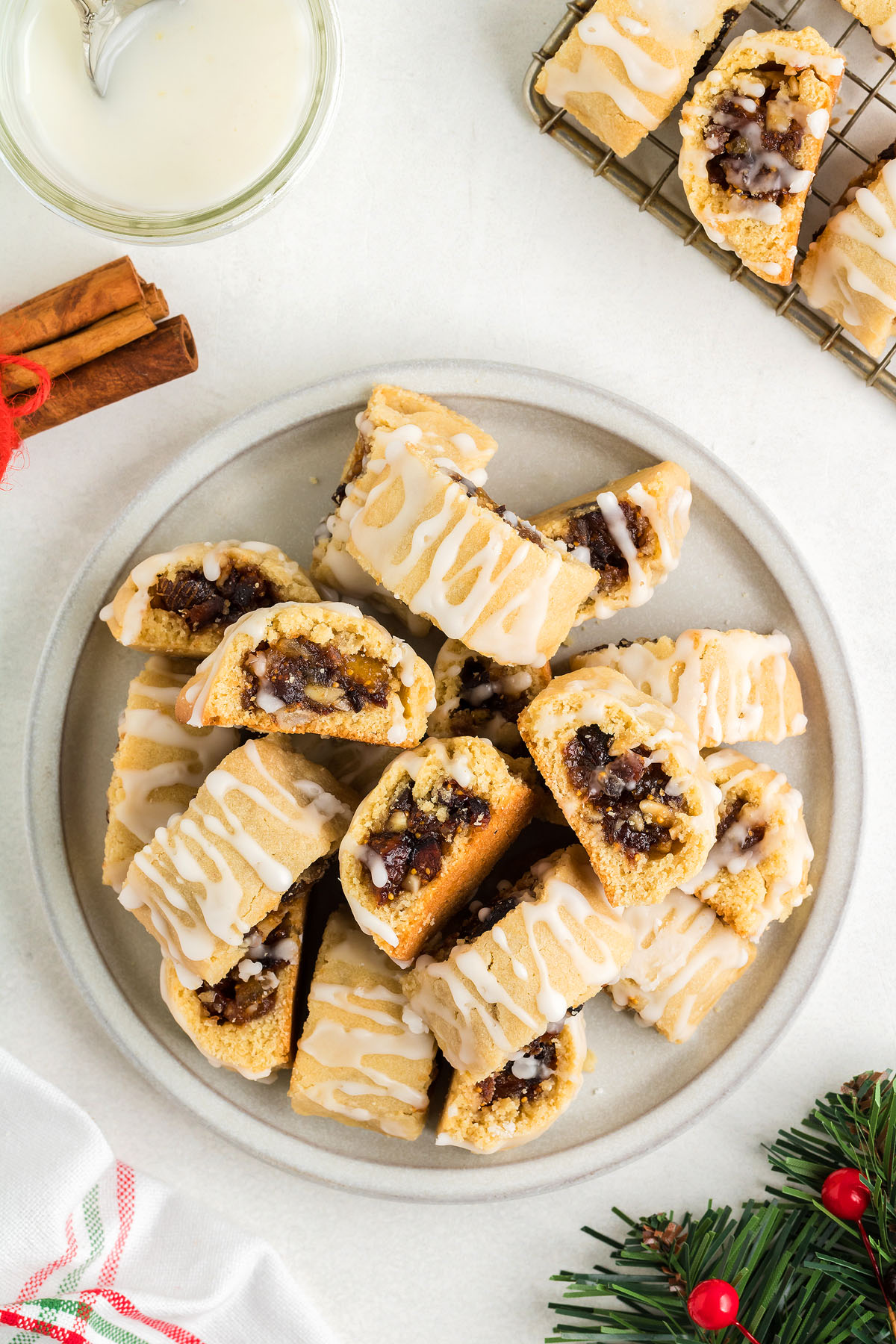 A plate of glazed, sliced Italian Fig Cookies – Cucidati with a spiced filling, surrounded by cinnamon sticks and holiday decor, brings classic Italian Christmas cookie charm to your table.