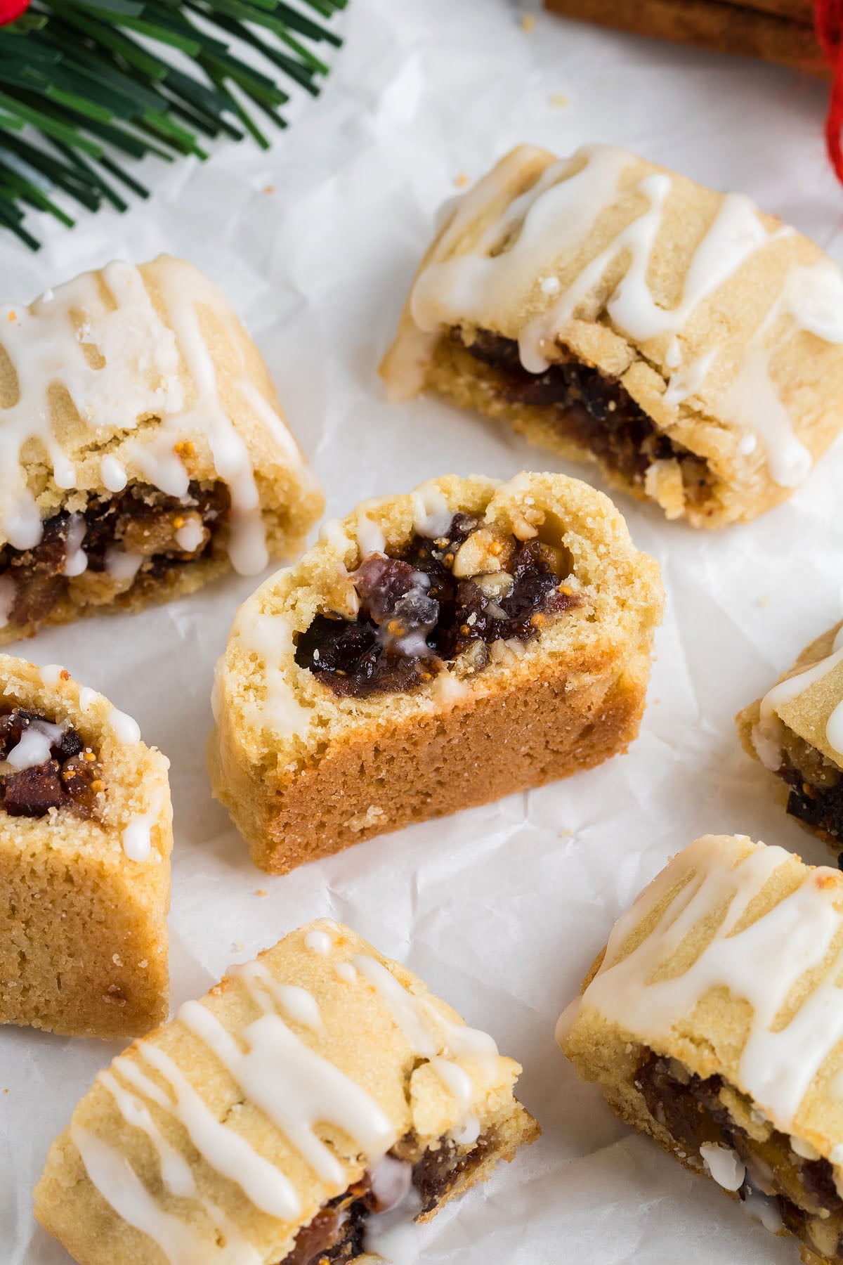 Italian Fig Cookies – Cucidati, with a sweet fruit filling and white icing drizzle, are arranged on parchment paper near pine tree branches, capturing the charm of traditional Italian Christmas cookies.