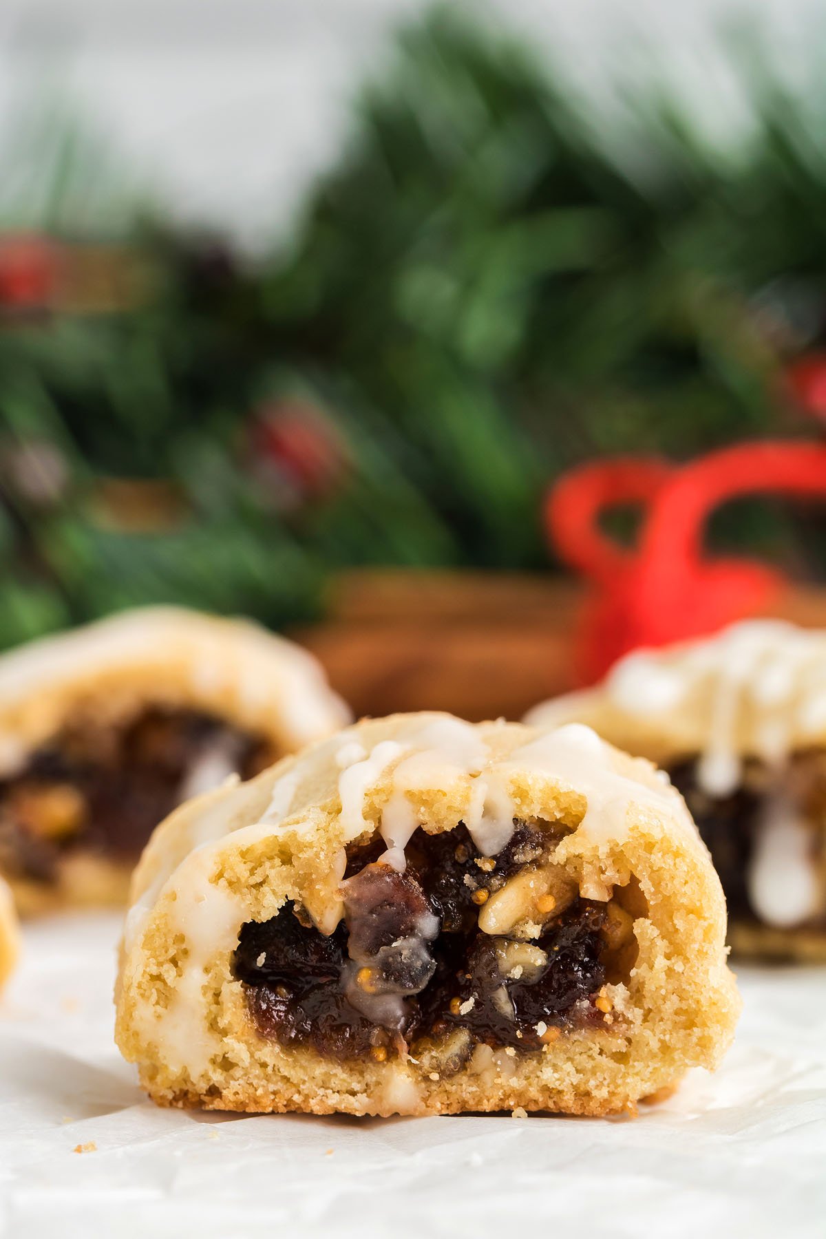Close-up of an Italian Fig Cookie – Cucidati, its glazed top and fruit and nut filling shining, with holiday greenery blurred in the background.