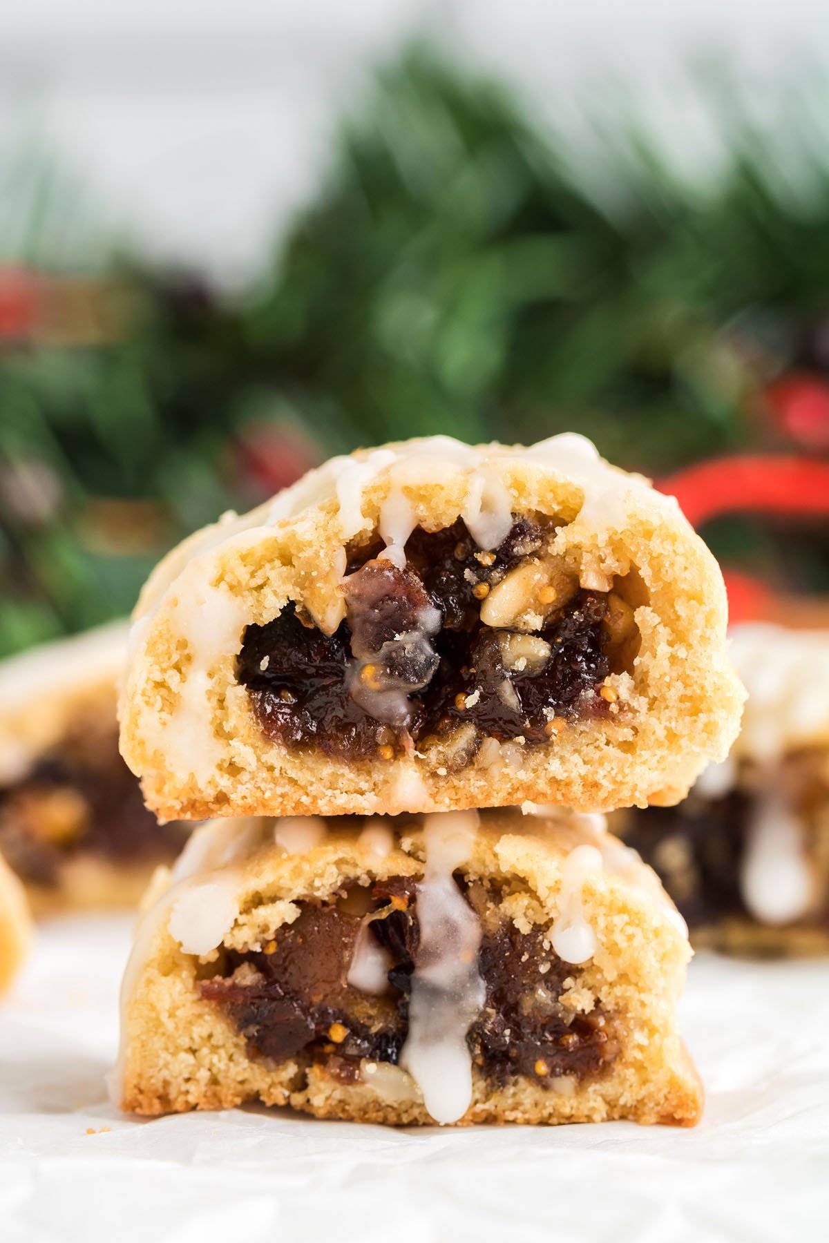 Two glazed Italian Fig Cookies – Cucidati, filled with fruit and nuts, are stacked in front of a festive, blurred holiday background.