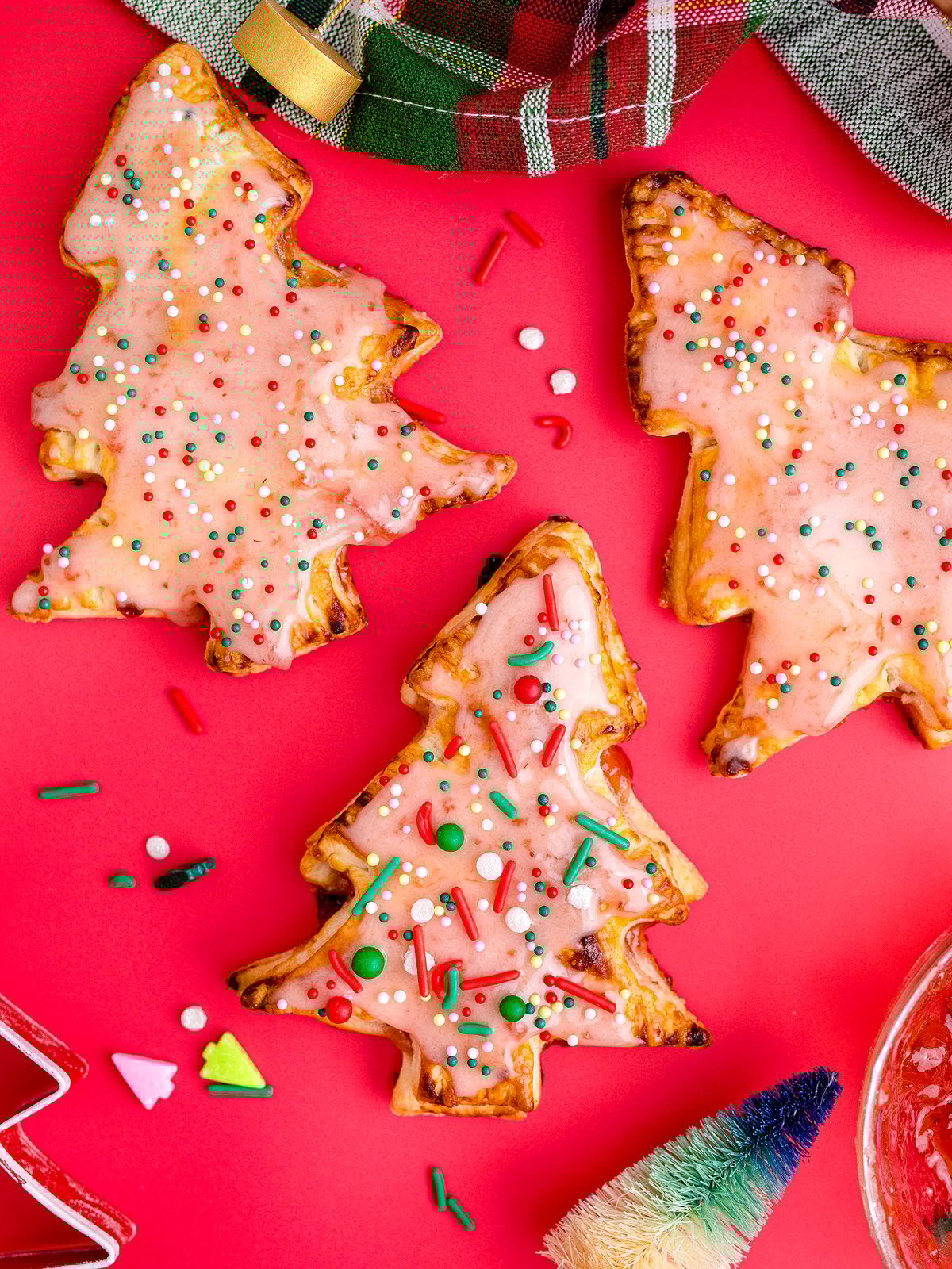 Three Christmas Tree Pop Tarts with pink icing and colorful sprinkles sit on a red background, making a festive treat inspired by holiday tart recipes.