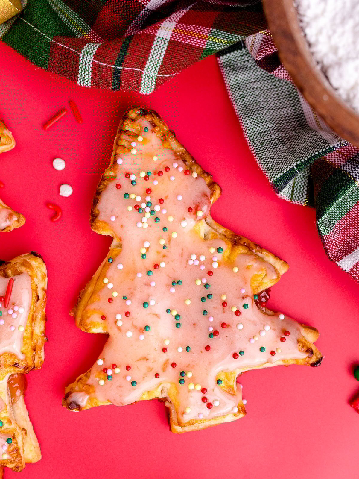 Christmas tree-shaped cookie with pink icing and colorful sprinkles on a red surface near a plaid cloth, inspired by festive Christmas Tree Pop Tarts for a delightful holiday treat.