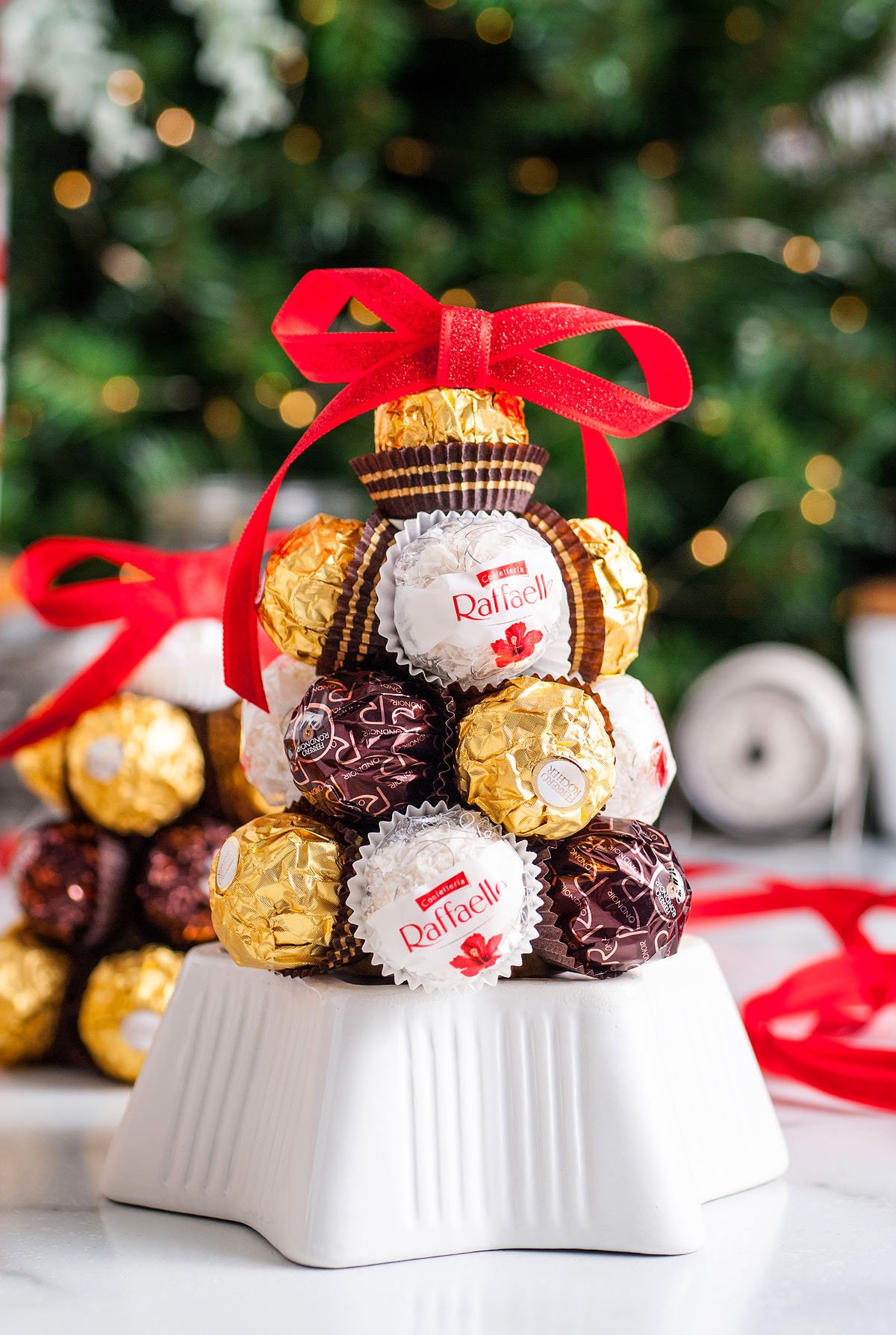 A pyramid of assorted Ferrero chocolates tied with a red ribbon sits in front of a decorated Christmas tree—an irresistible Homemade Ferrero Rocher Christmas Tree perfect as a festive DIY Christmas decoration or a thoughtful Christmas hostess gift.