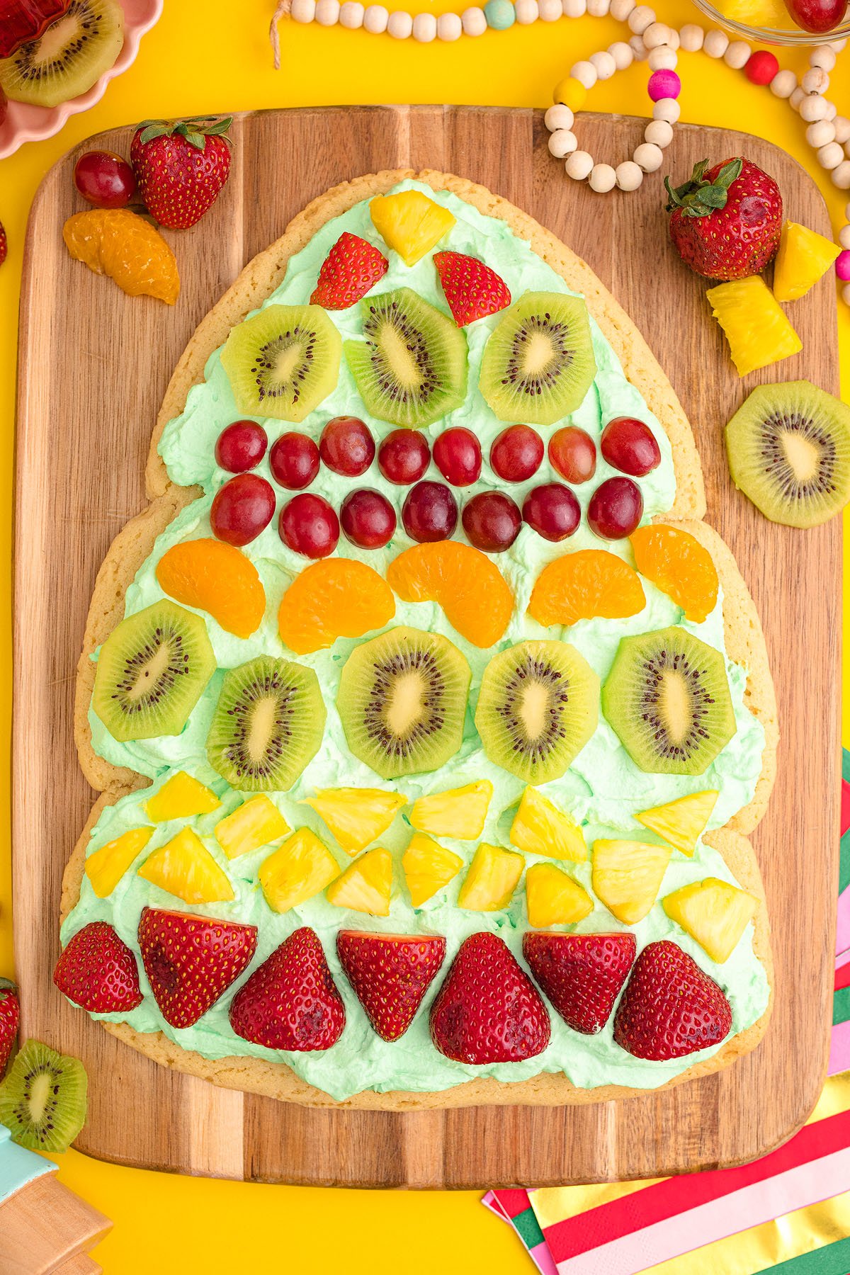 A tree-shaped Christmas Tree Fruit Sugar Cookie dessert pizza with green frosting, topped with strawberries, kiwi, grapes, oranges, and pineapple—perfect as a festive Christmas dessert!.