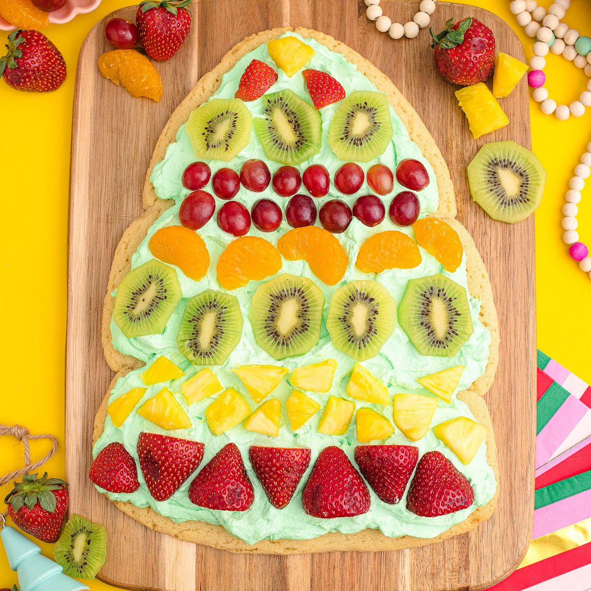 A large, tree-shaped fruit pizza reminiscent of a Christmas Tree Fruit Sugar Cookie, topped with strawberries, kiwi, pineapple, grapes, and orange slices—perfect as a festive Christmas dessert.