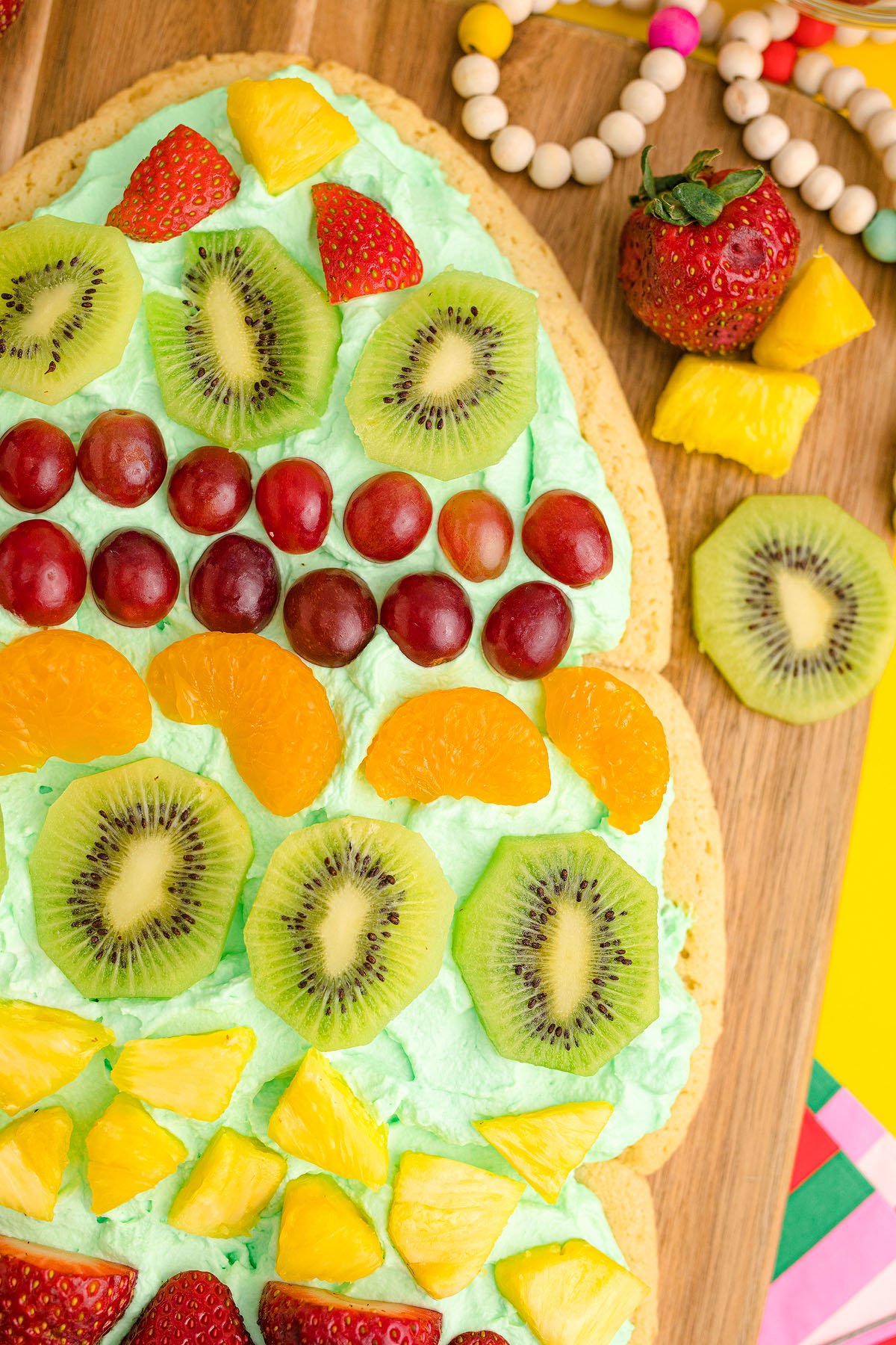 Easter egg-shaped fruit pizza with green frosting, topped with kiwi, strawberries, grapes, oranges, and pineapple—a festive twist similar to a Christmas Tree Fruit Sugar Cookie.