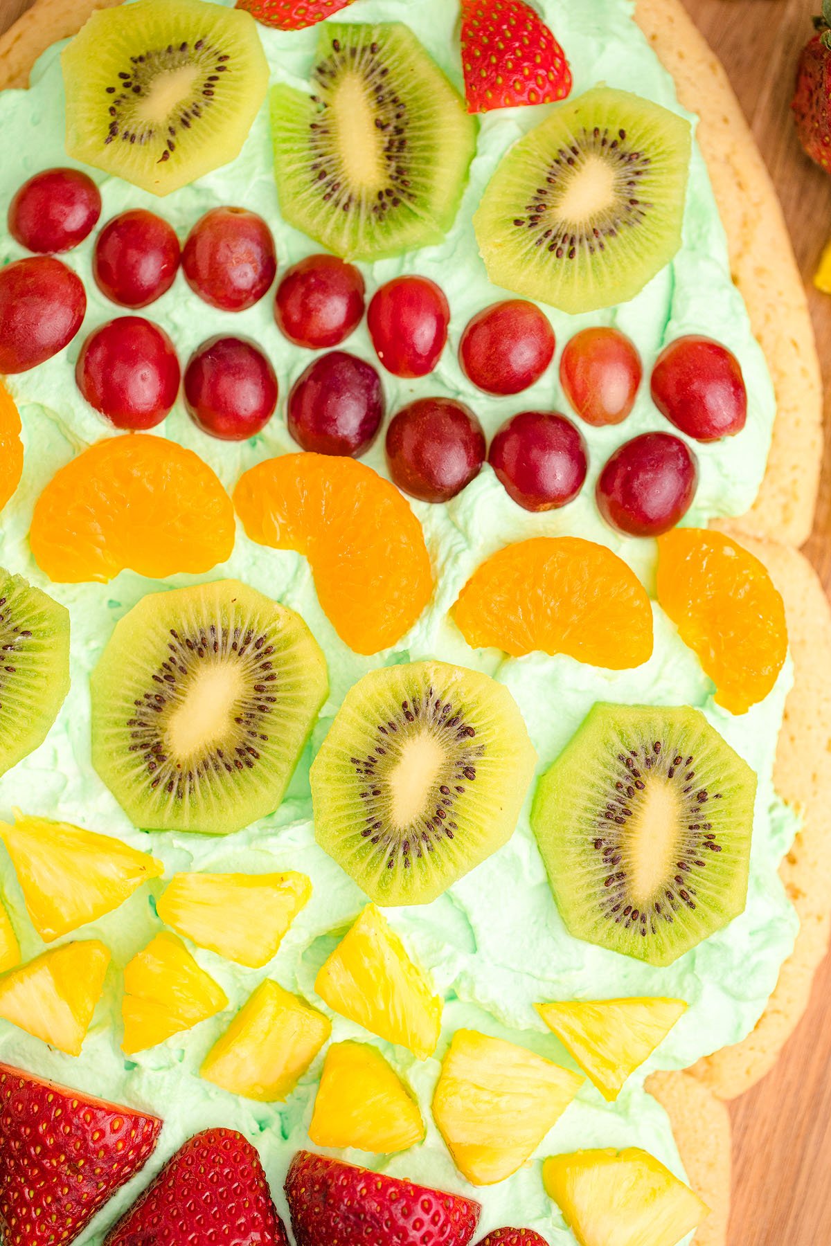 A close-up of a tree shaped fruit pizza, perfect as a Christmas dessert, topped with kiwi, grapes, mandarin oranges, pineapple, and strawberries.