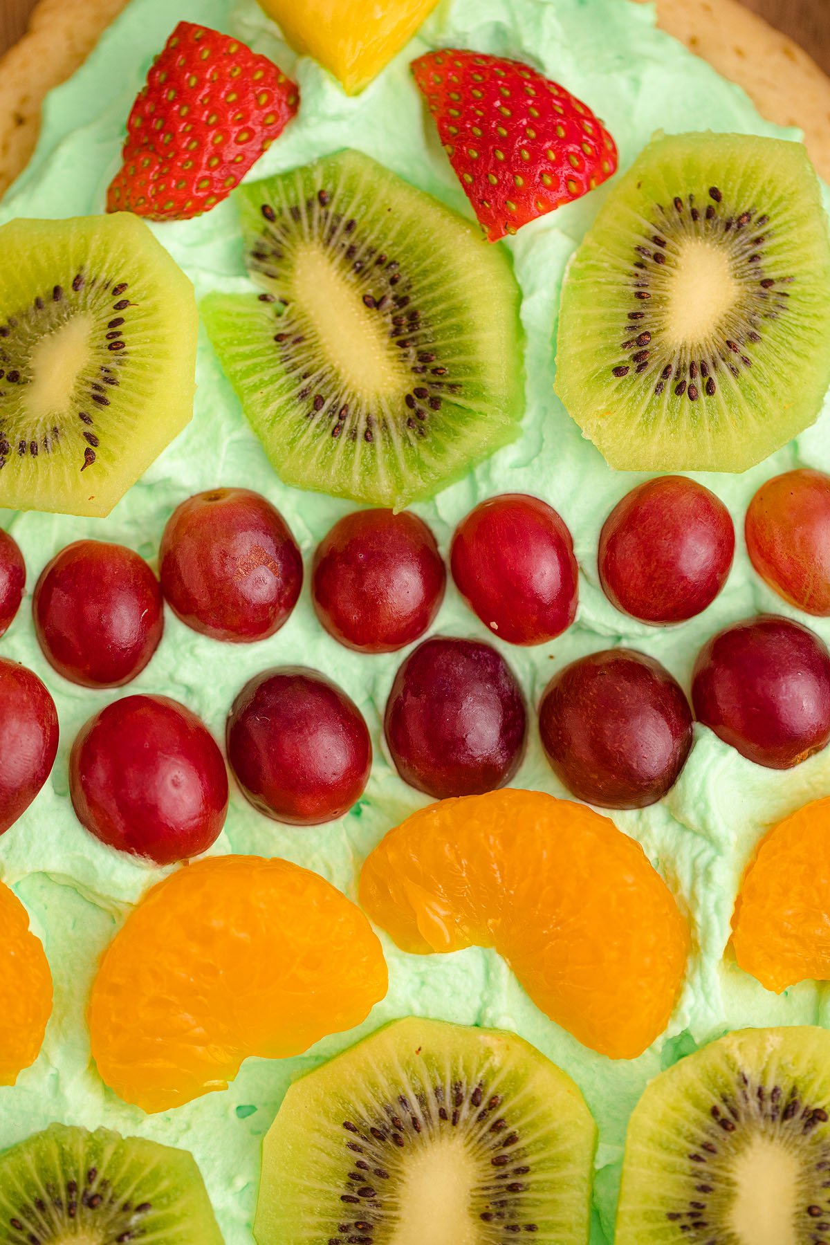 Close-up of a tree shaped Christmas Tree Fruit Sugar Cookie, topped with sliced kiwi, strawberries, grapes, and orange segments on green whipped cream—perfect as a festive Christmas dessert.