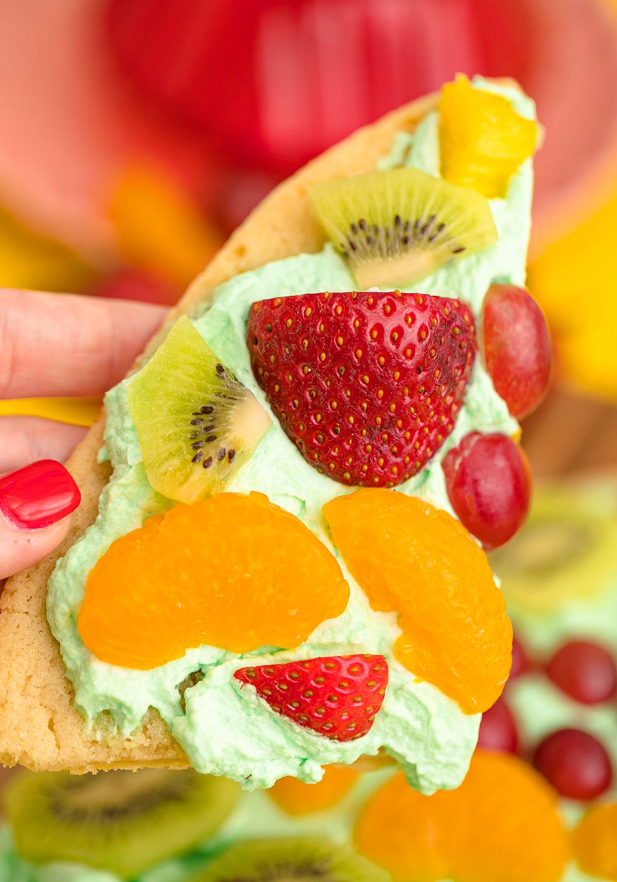 A hand holding a festive Christmas Tree Fruit Sugar Cookie pizza topped with green cream, strawberries, kiwi, grapes, and orange segments—perfect as a colorful Christmas dessert.