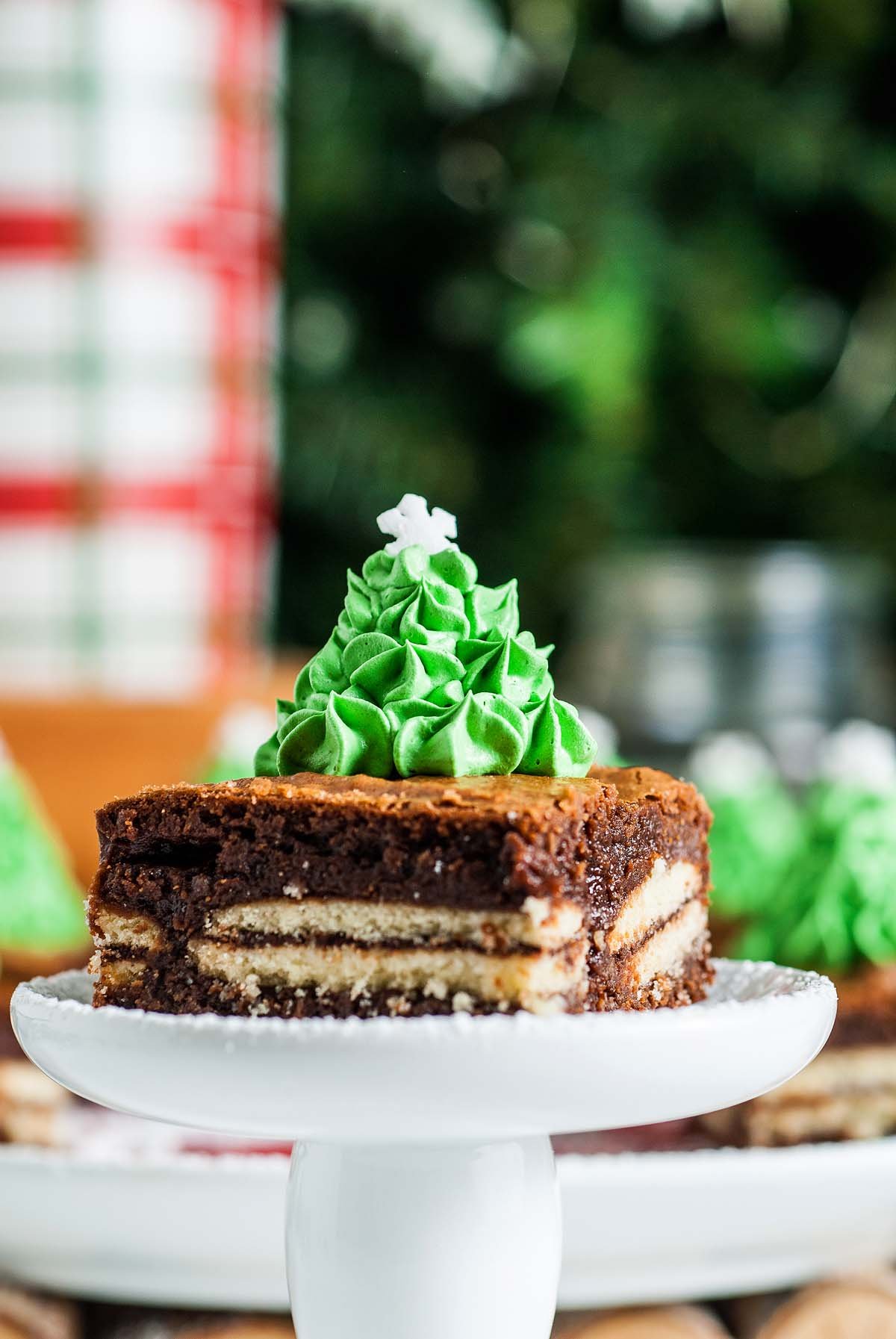 A Mint Milano Christmas Brownie topped with green icing, decorated with a Christmas tree, sits on a white cake stand.