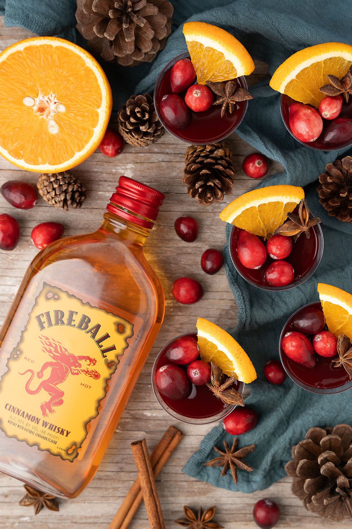 Fireball whisky, Mulled Wine Fireball Jello Shots, cocktails with orange slices and cranberries, pinecones, and cinnamon sticks on a wooden table.