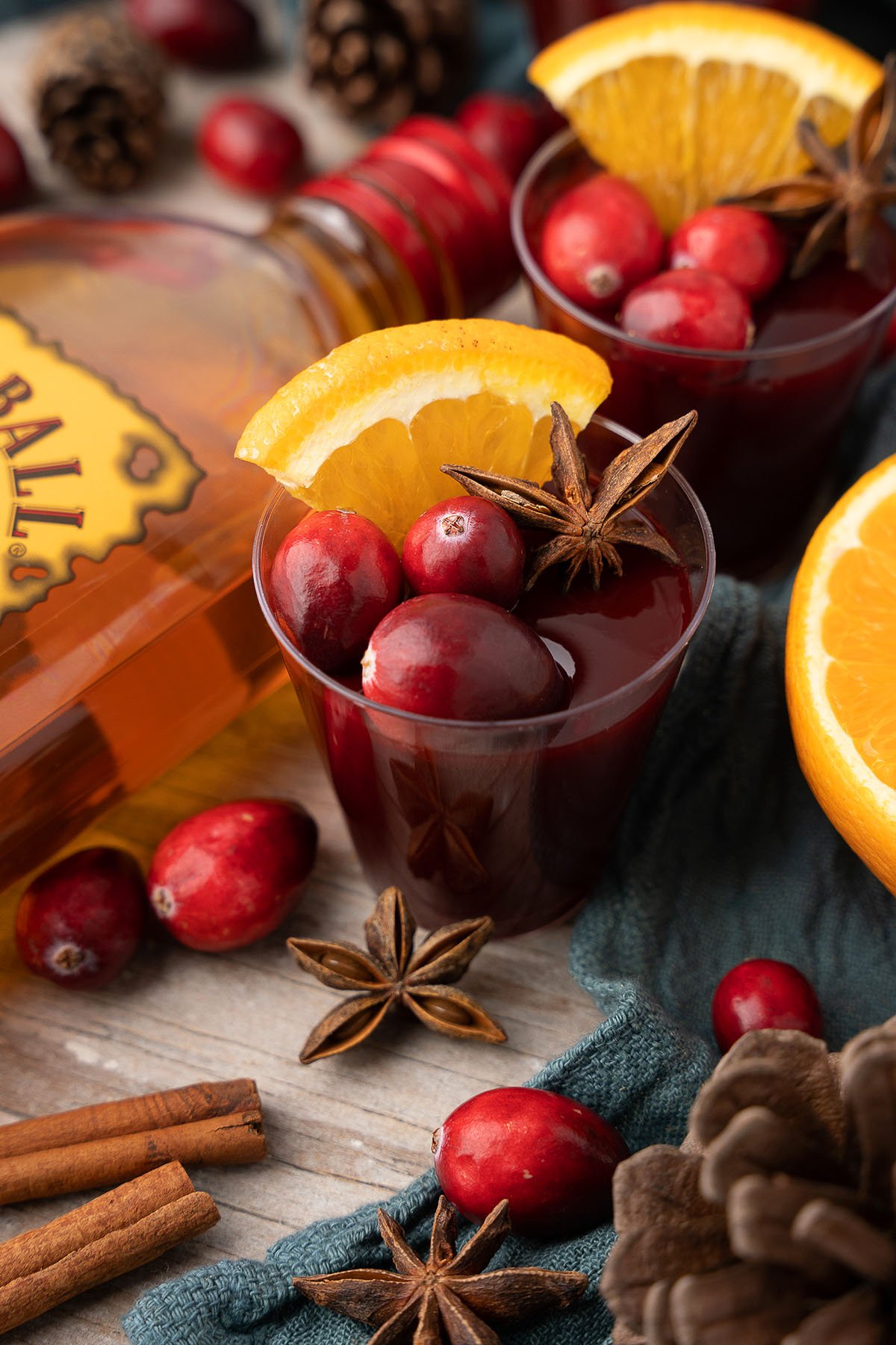 A cup of mulled wine garnished with cranberries, orange slice, and star anise sits nearby a bottle and spices—perfect inspiration for festive drinks like Mulled Wine Fireball Jello Shots or other Christmas shot recipes.