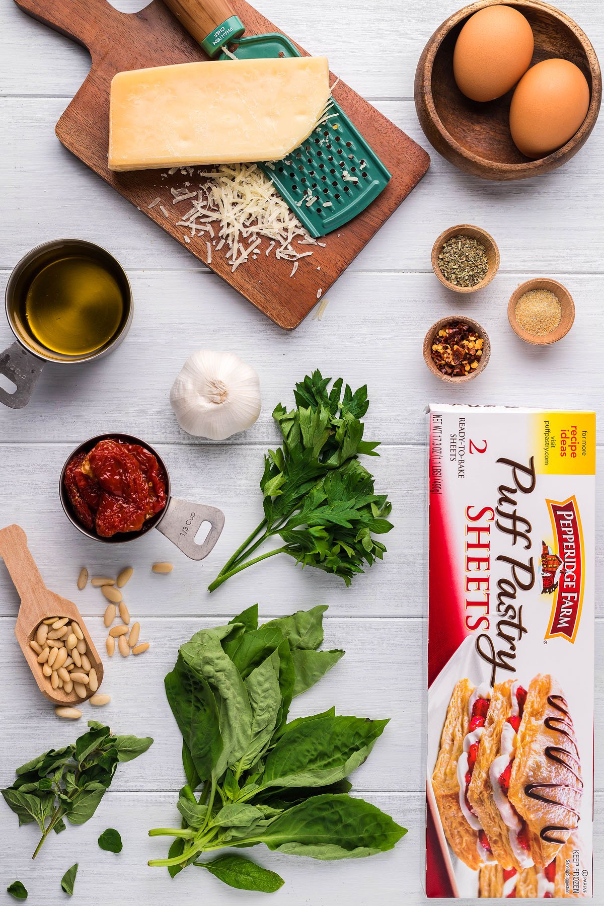 Ingredients for a festive puff pastry recipe, including eggs, cheese, herbs, olive oil, and seasonings on a white surface—perfect for making Pesto Puff Pastry Christmas Trees or other delicious Christmas appetizers.