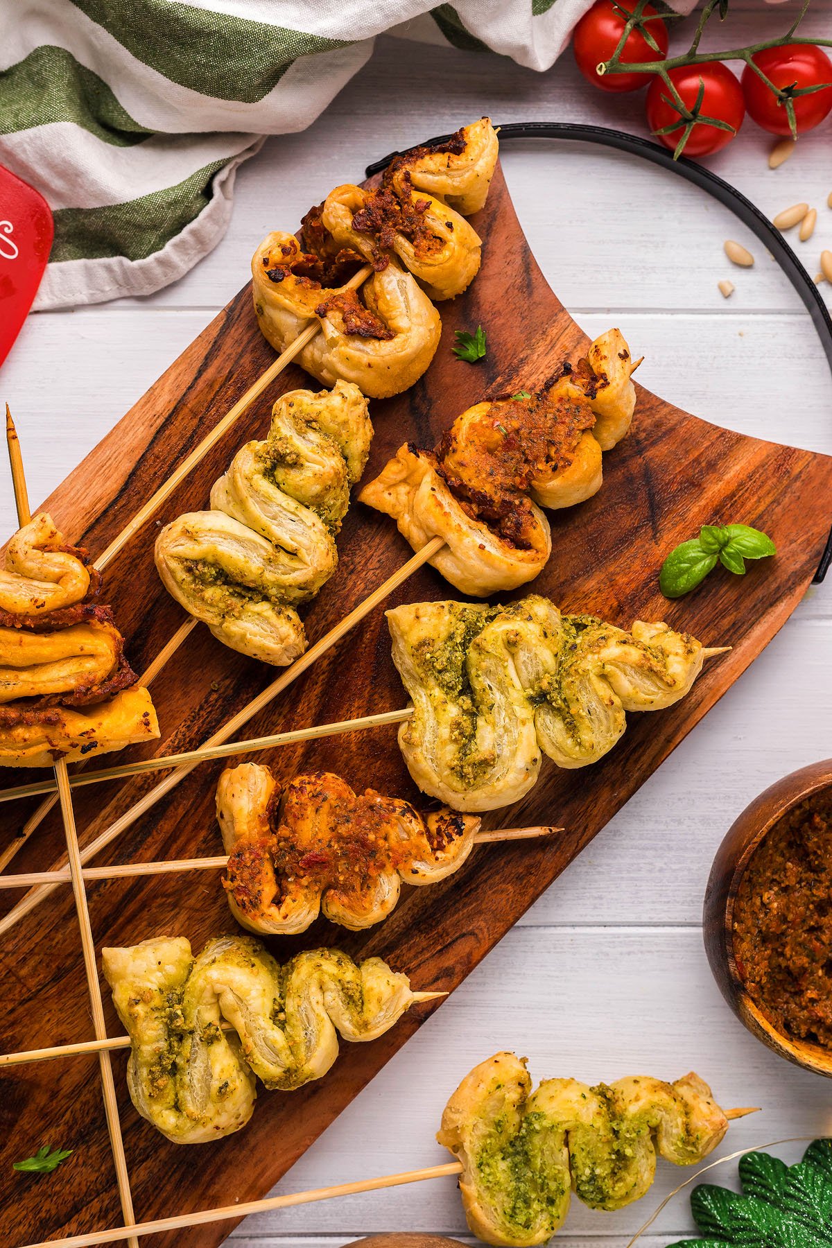 Skewered puff pastry twists with pesto and tomato spreads on a wooden board, surrounded by fresh herbs—these Pesto Puff Pastry Christmas Trees make festive and delicious Christmas appetizers.